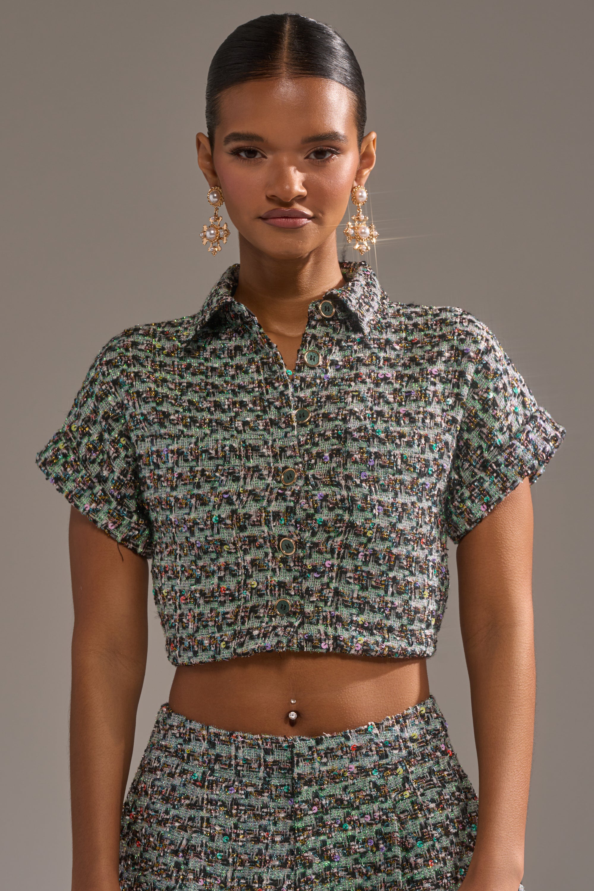 A woman with slicked-back hair wears the BRIGETTE TWEED BUTTON DOWN as a cropped, textured, multicolored top with matching high-waisted pants, accessorized with dangling earrings and a belly button piercing against a neutral background. 0