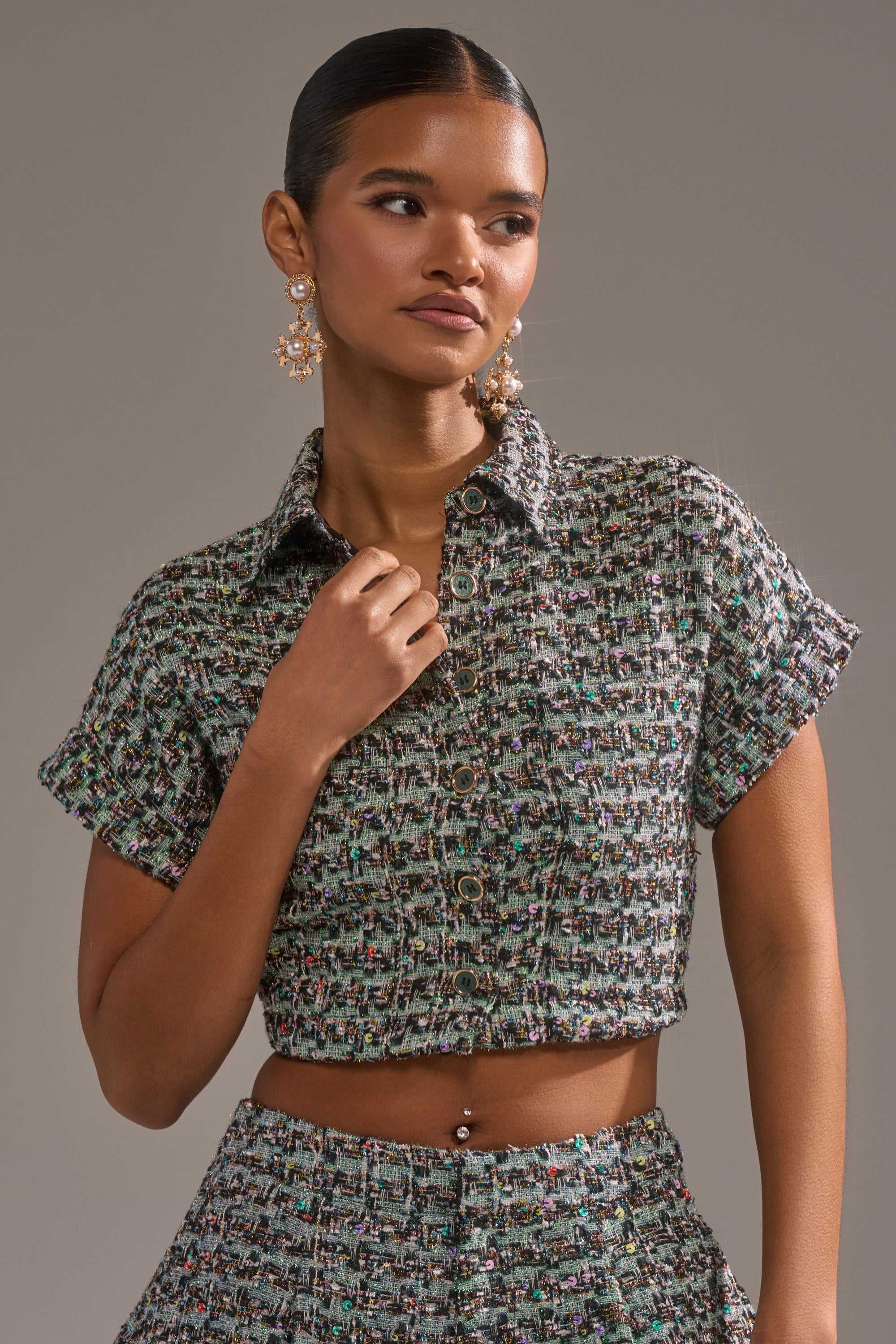 A woman with slicked-back hair and statement earrings poses confidently in the BRIGETTE TWEED BUTTON DOWN and matching high-waisted pants, one hand near her collar as she looks to the side. 1