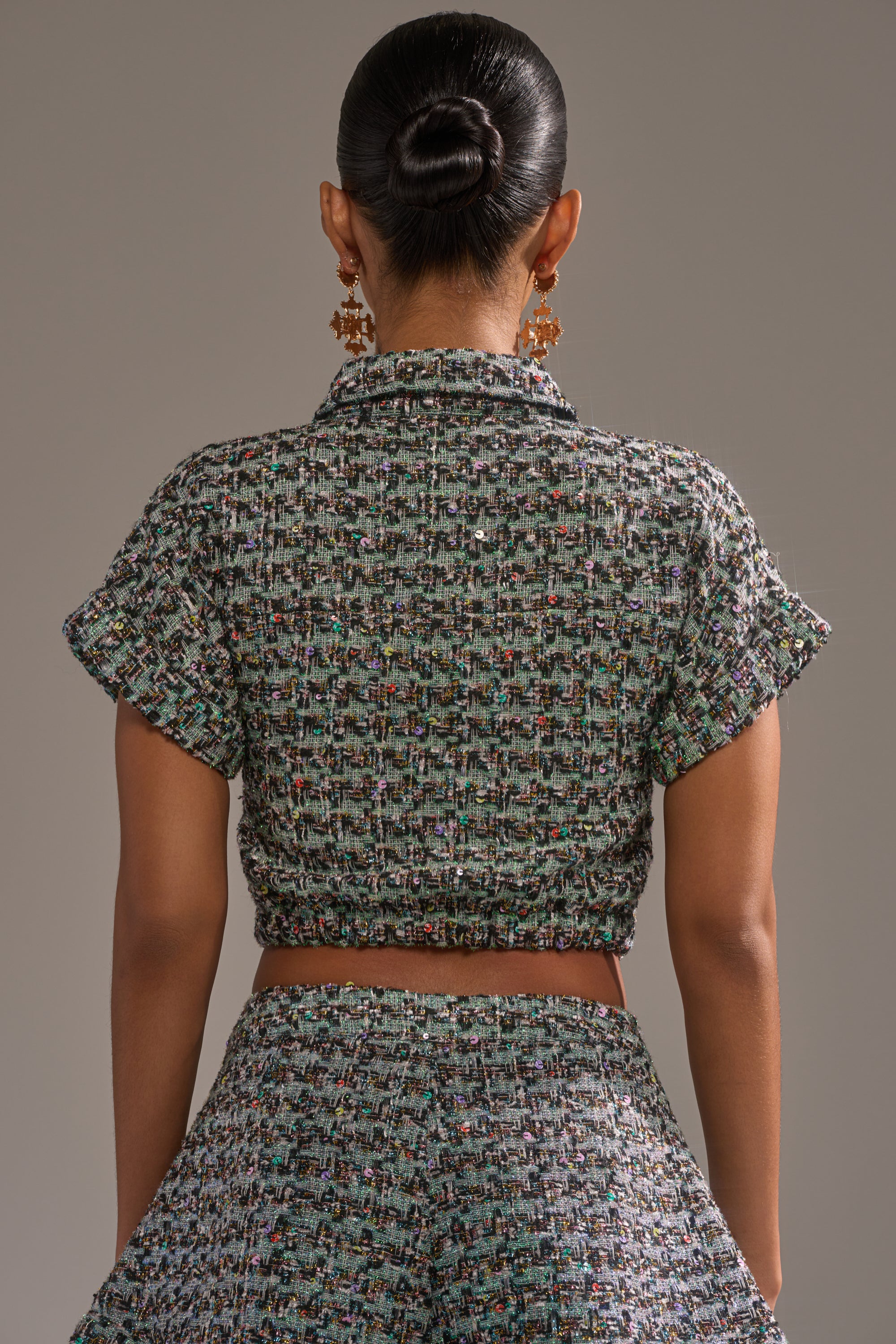 A woman faces away, wearing the BRIGETTE TWEED BUTTON DOWN— a cropped metallic tweed top with a matching high-waisted skirt. Her sleek bun and large statement earrings complete the look. 3