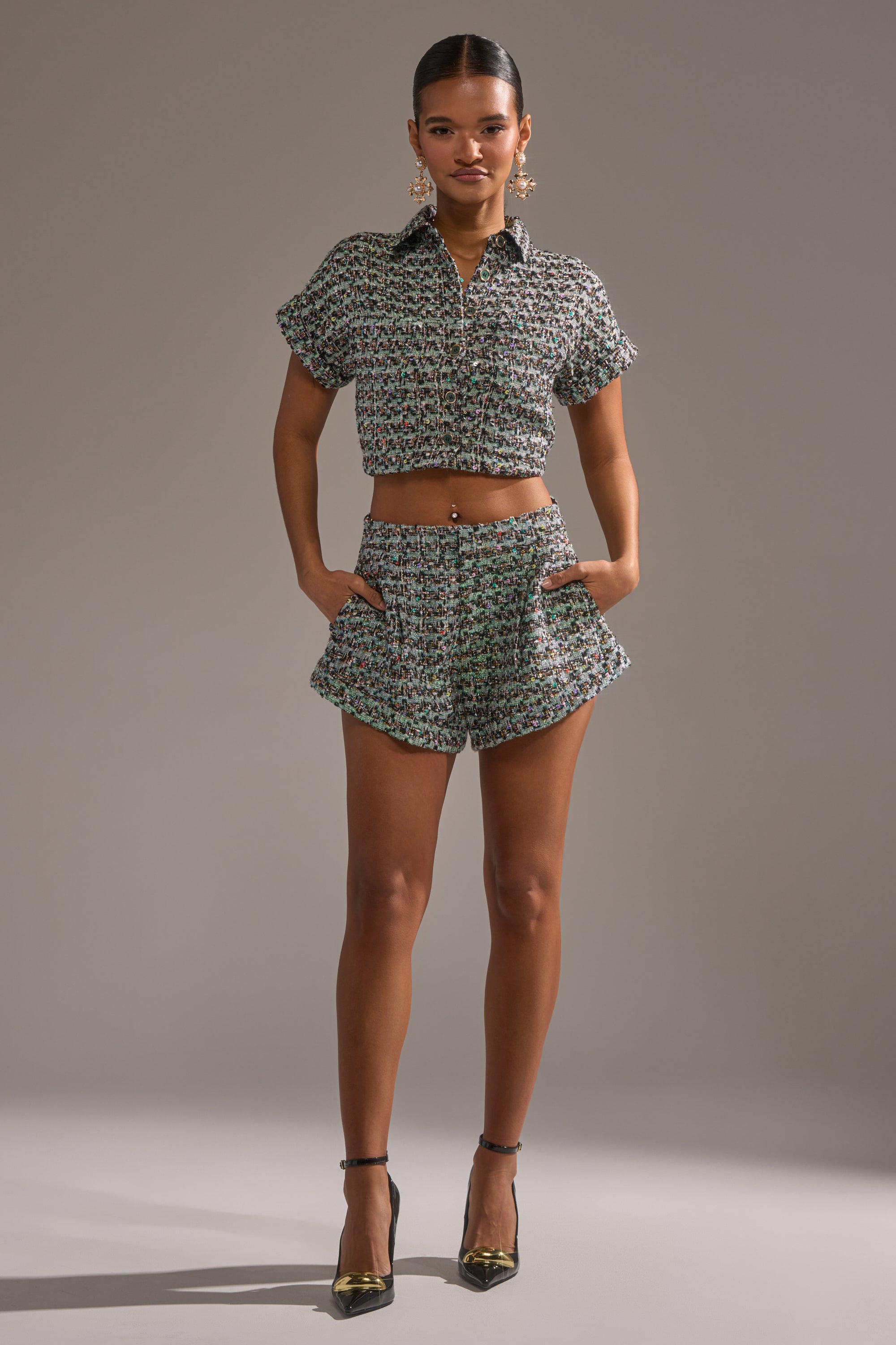 A woman faces forward in the BRIGETTE TWEED BUTTON DOWN, a matching textured short-sleeve cropped button down and high-waisted shorts set, paired with black heels, gold earrings, and a neat ponytail against a plain background. 4
