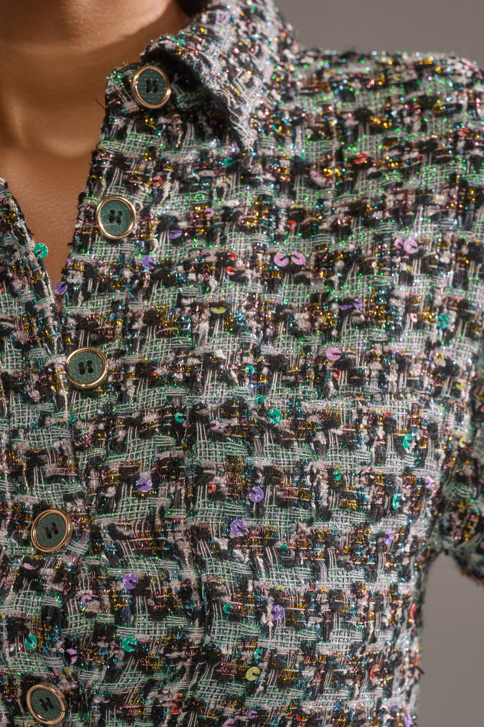 Close-up of the BRIGETTE TWEED BUTTON DOWN, a cropped shirt in textured, multicolored tweed with metallic threads, sequins, and green buttons. Partially buttoned and modeled to show upper chest and shoulder. 5
