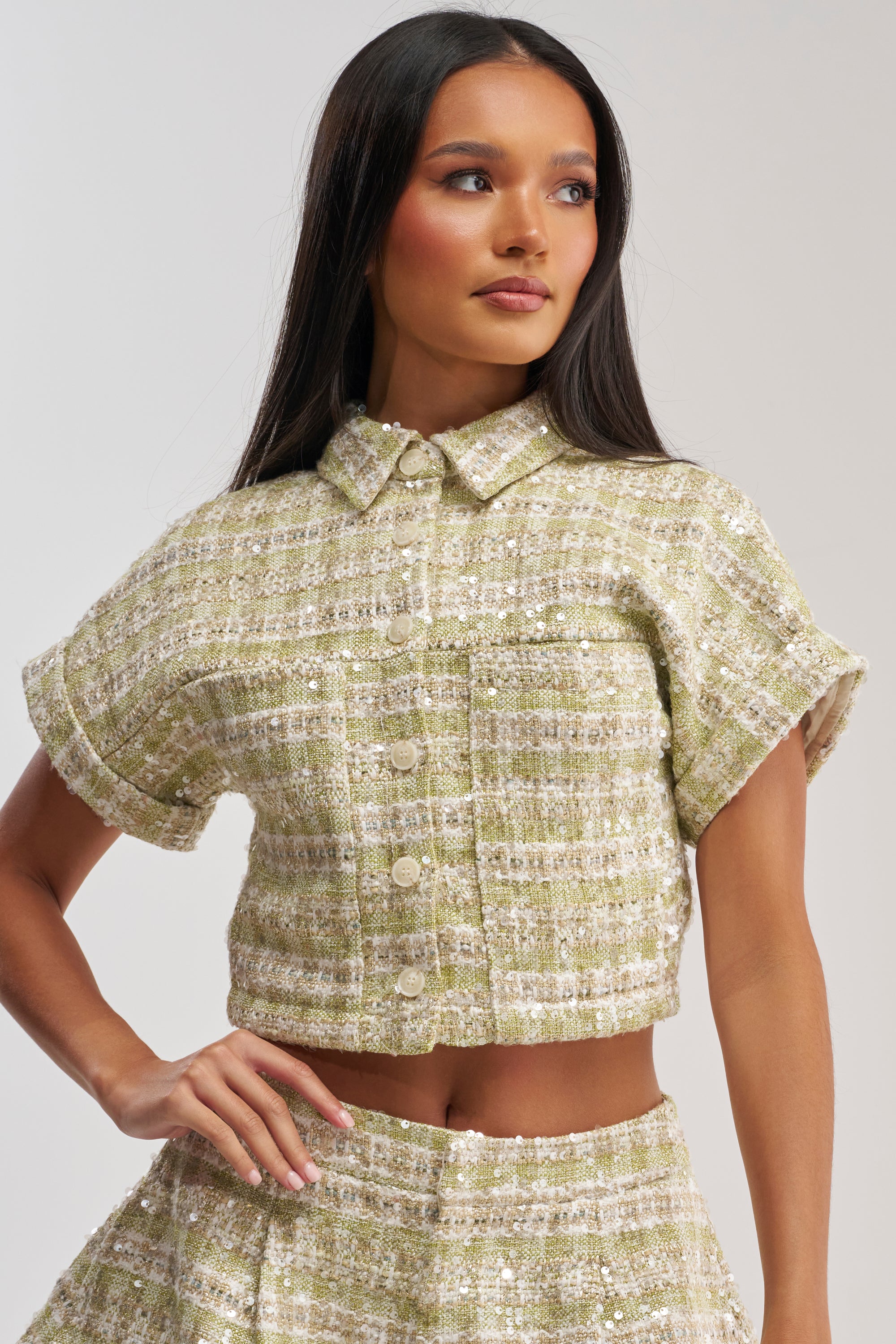 A woman exudes confidence in the EASY TO LOVE TWEED BUTTON DOWN, a short-sleeved, collared cropped blouse paired with a matching green and white checkered tweed skirt—perfect for spring brunches. Her long, straight hair enhances the chic vibe.