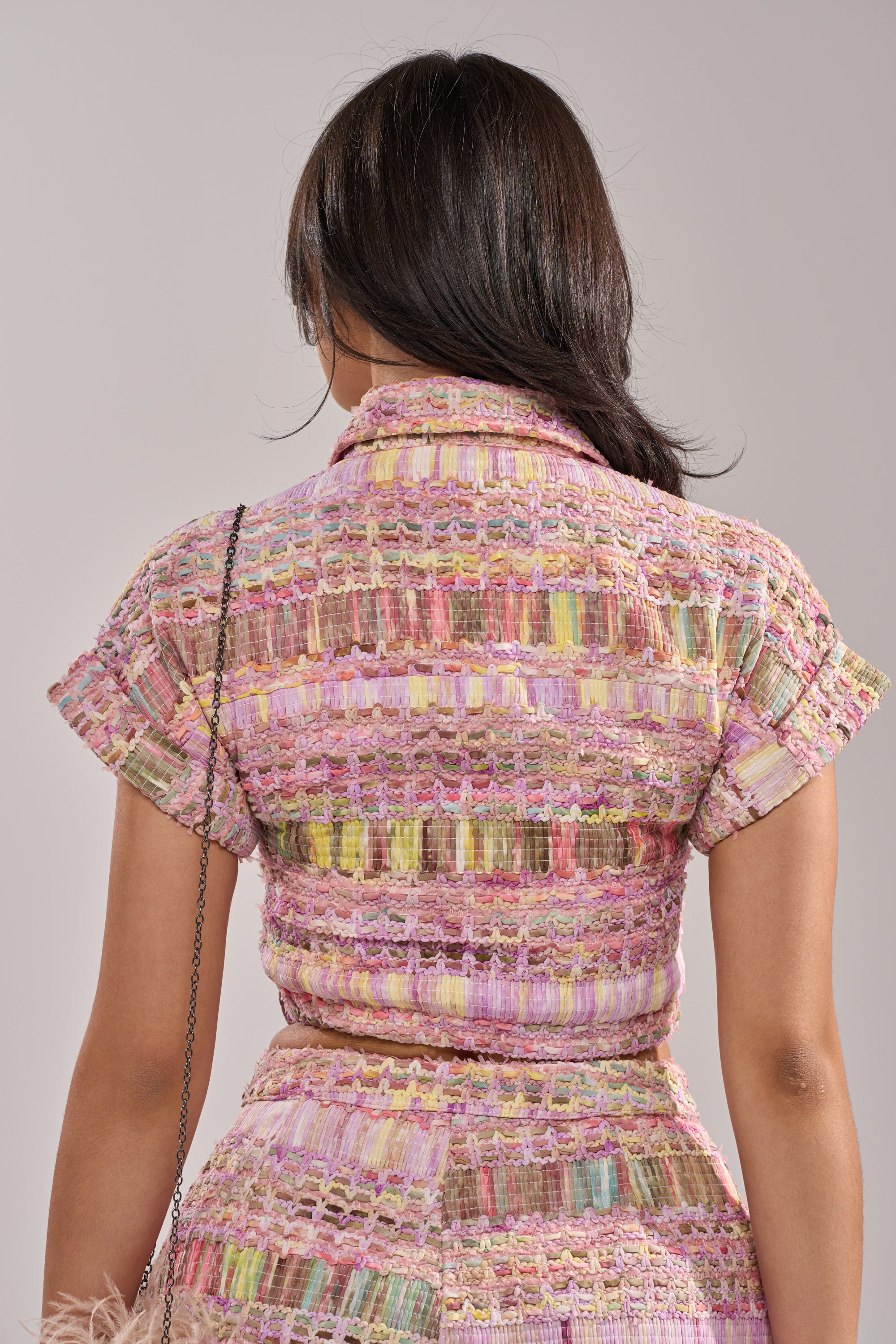 A woman with medium-length dark hair is seen from behind wearing the VERONICA TWEED PLAID BLOUSE in pastel plaid, paired with a matching skirt. She accessorizes with a small chain-strap bag on her shoulder. 4