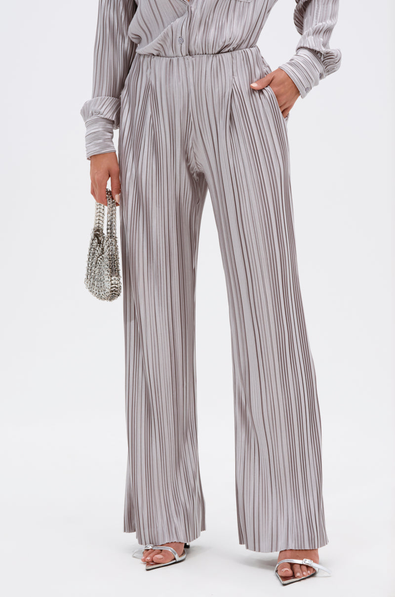 A person wears the ALIA SLINKY PALAZZO PANT in light gray satin-blend with accordion pleats, paired with a matching long-sleeve top, white heeled sandals, and a silver beaded purse. The setting is a plain white background. 1