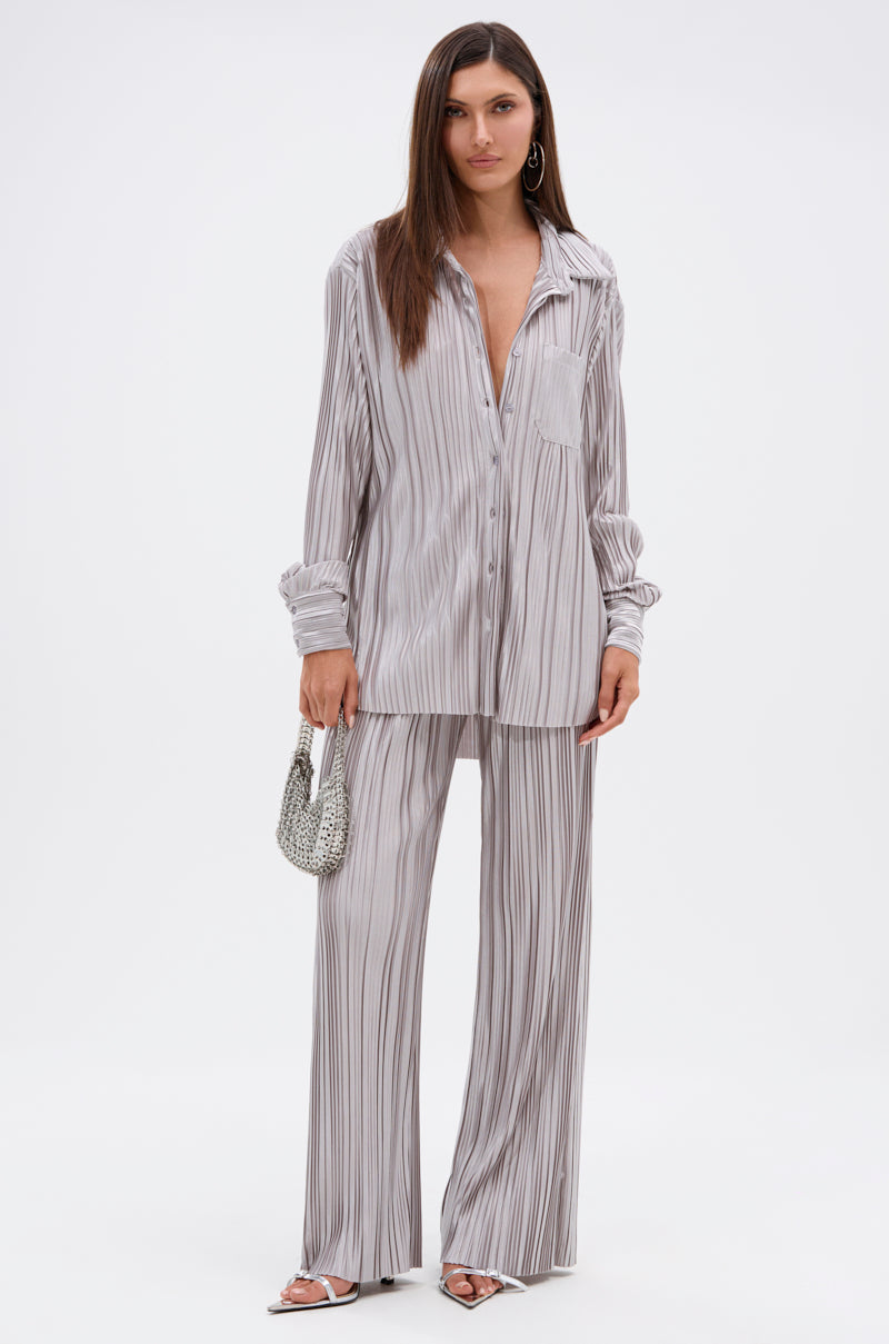 A woman with long straight hair and hoop earrings stands against a white background, wearing the ALIA SLINKY PALAZZO PANT and a loose-fit light gray striped shirt. She accessorizes with sandals and a small textured handbag. 5