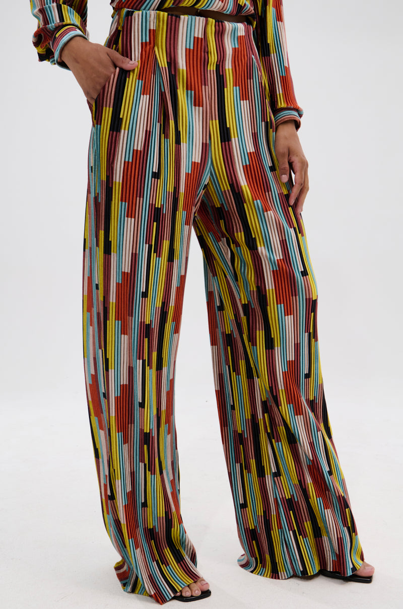 Wearing the VIBES FEEL GOOD VELVET PALAZZO PANT, featuring a colorful vertical striped pattern in red, yellow, black, blue, and white; one hand is in the pocket and only the lower half of the body is visible. 2
