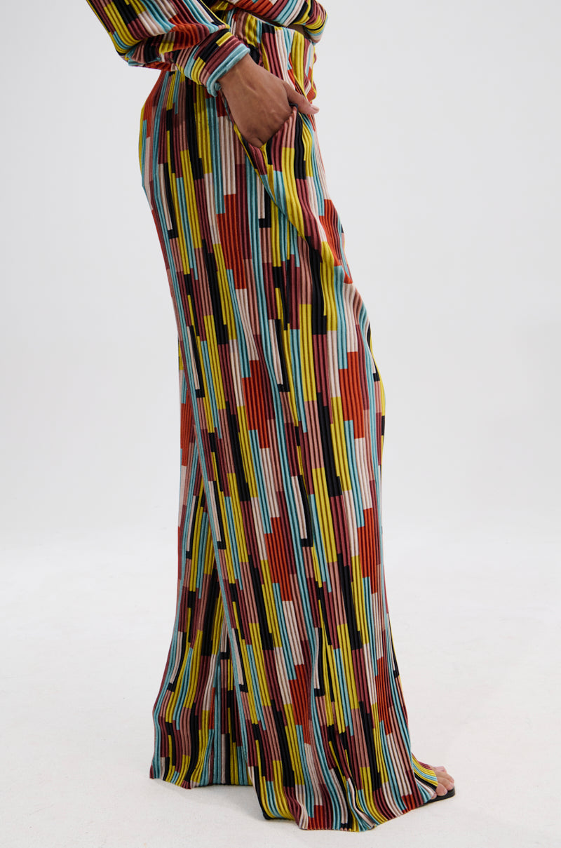 A person stands barefoot in VIBES FEEL GOOD VELVET PALAZZO PANT, featuring a wide-leg silhouette and colorful geometric stripes in red, yellow, blue, black, and white. A matching top sleeve is partially visible against a plain white background. 3