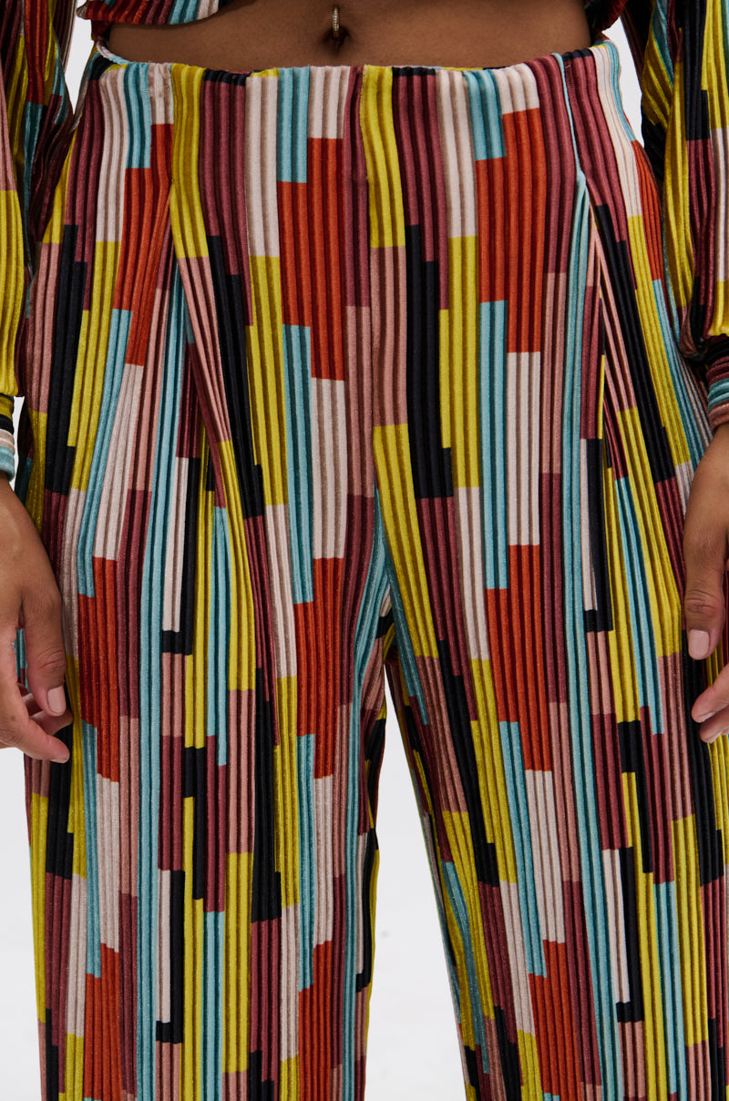 A close-up of a person wearing the VIBES FEEL GOOD VELVET PALAZZO PANT, featuring a high-waisted fit and colorful geometric pattern with vertical stripes in red, yellow, blue, black, and pink. The wide-leg silhouette is shown with hands at the sides. 7