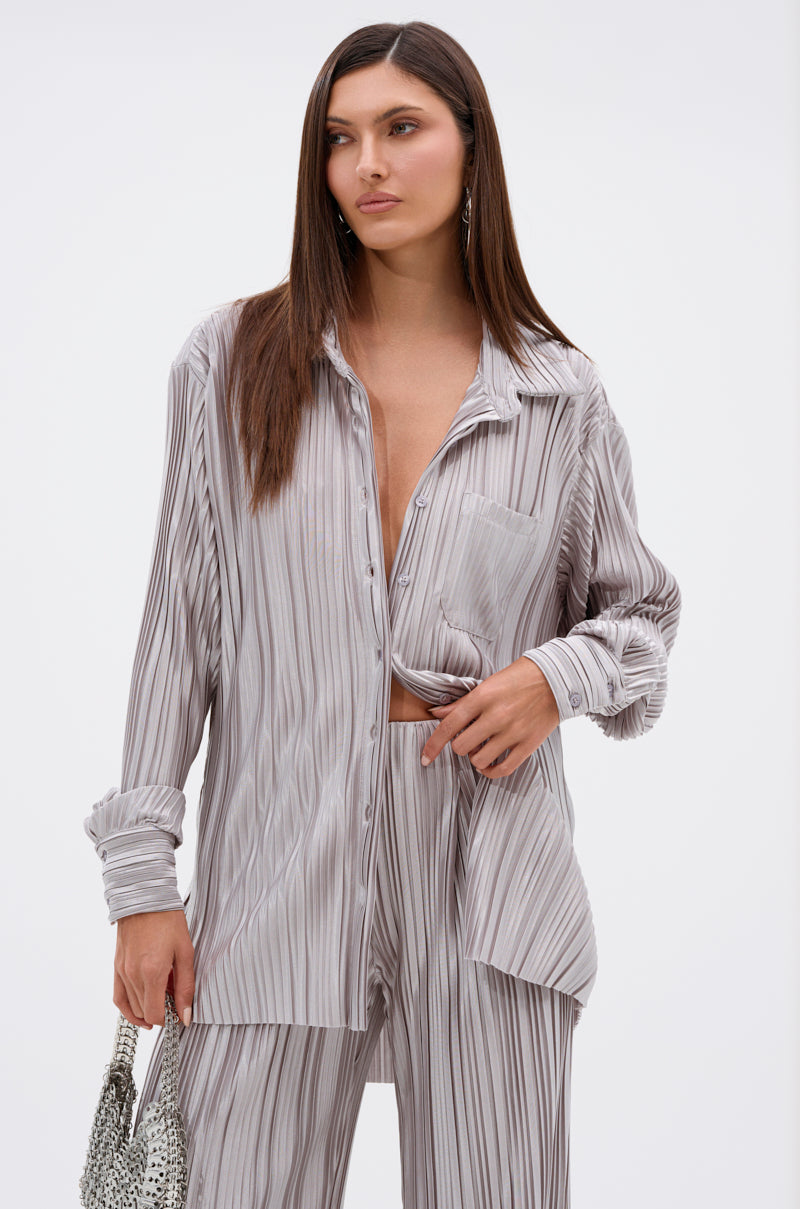 A woman poses against a plain white background, wearing the ALIA SLINKY BUTTON DOWN—a loose-fitting, light gray, pinstriped satin-blend shirt with matching trousers—and holds a silver bag with one hand on her waist.