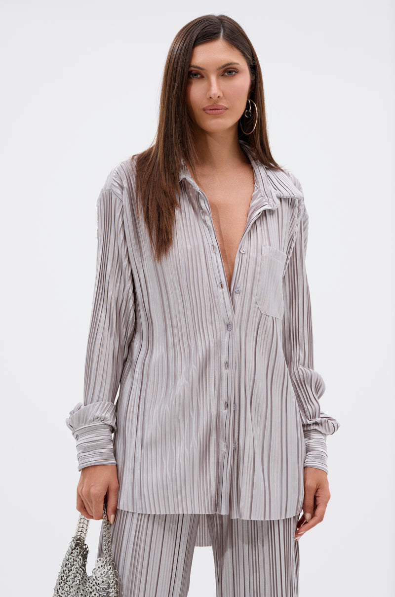 A woman in the ALIA SLINKY BUTTON DOWN, a shimmering silver shirt paired with matching pants, stands against a plain background holding a textured handbag in her left hand.