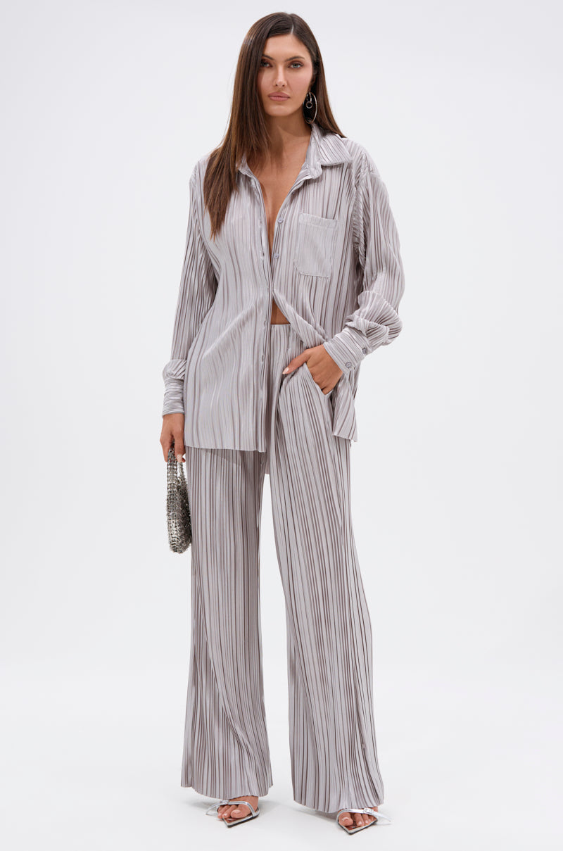A woman stands against a white background wearing the ALIA SLINKY BUTTON DOWN in shimmering silver pinstripes with matching wide-leg pants. She holds a small handbag, wears heels, lets her hair down, and has one hand in her pocket.