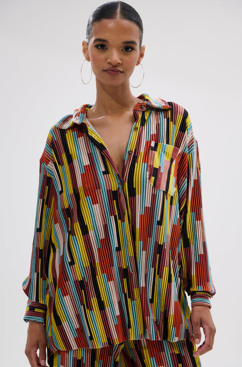 A woman with hoop earrings wears the VIBES FEEL GOOD VELVET COLOR BLOCKED BUTTON DOWN, a colorful oversized shirt with velvet stripes in red, yellow, blue, black, and white. It features a front pocket and collar. She faces forward with a slight smile. 0