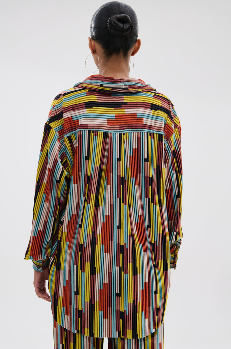 A person with dark hair in a bun is seen from behind, wearing the VIBES FEEL GOOD VELVET COLOR BLOCKED BUTTON DOWN—a colorful, retro-inspired shirt with geometric stripes in red, yellow, black, blue, and white. 2