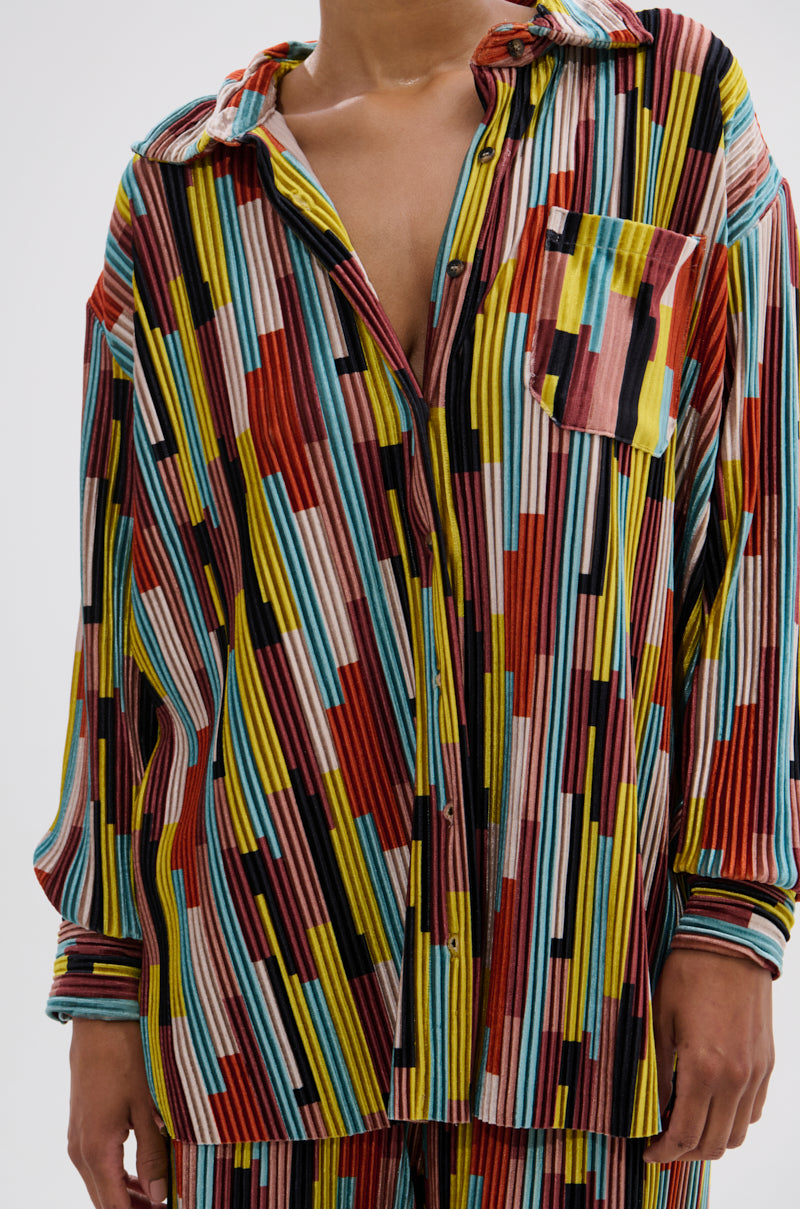 A person wears the VIBES FEEL GOOD VELVET COLOR BLOCKED BUTTON DOWN, a loose retro blouse with colorful vertical and horizontal stripes in red, yellow, teal, white, and black, partially unbuttoned and paired with matching pants. 6