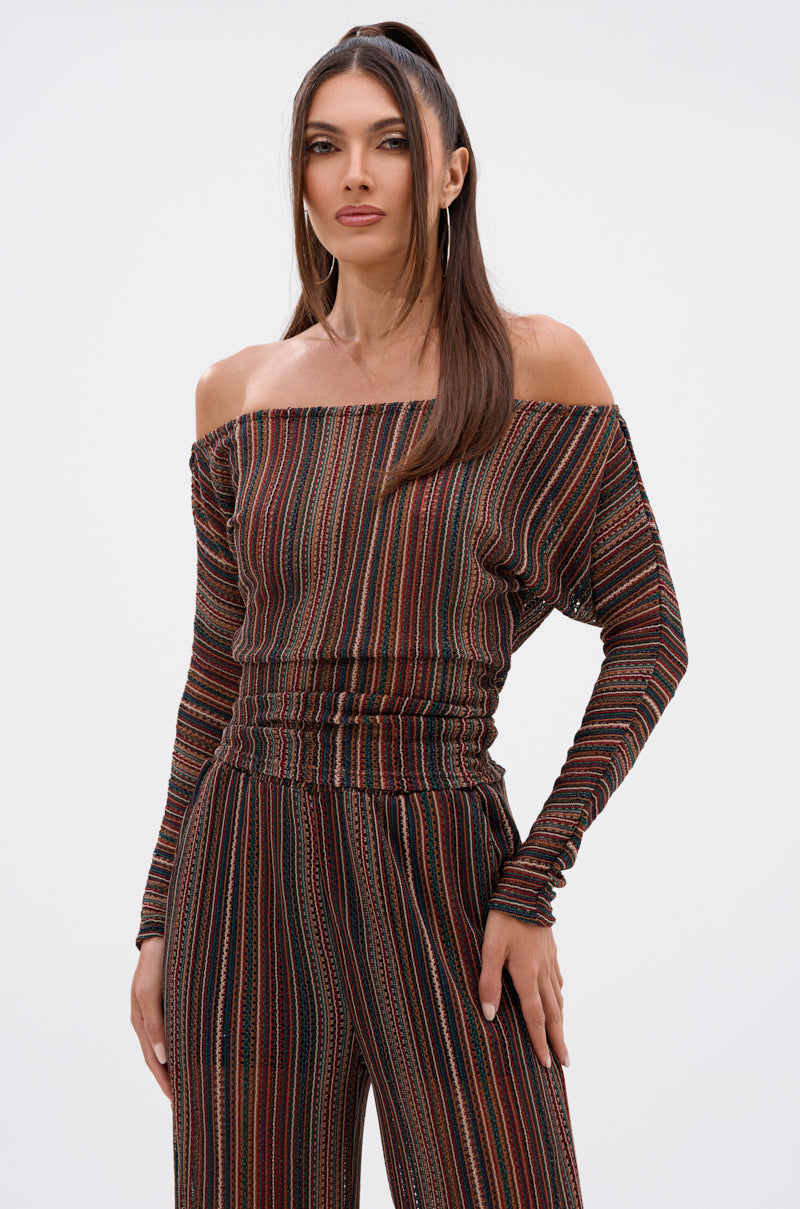 A woman with long, straight hair wears the CANDICE STRIPE OFF THE SHOULDER BLOUSE, styled as a striped jumpsuit in earthy tones. She stands facing forward with a neutral expression against a plain white background. 0
