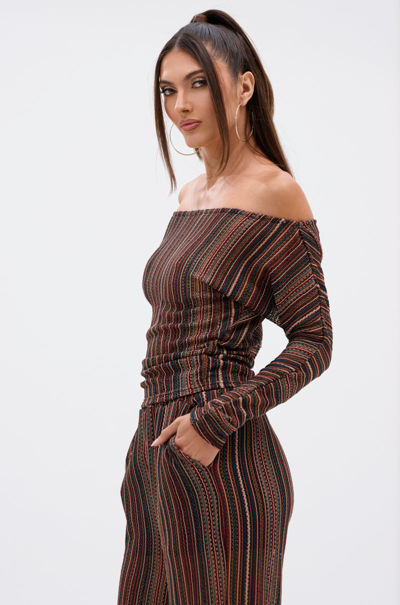 A woman with long brown hair in a high ponytail wears the CANDICE STRIPE OFF THE SHOULDER BLOUSE and matching pants, her hand in her pocket, standing against a plain white background. 2