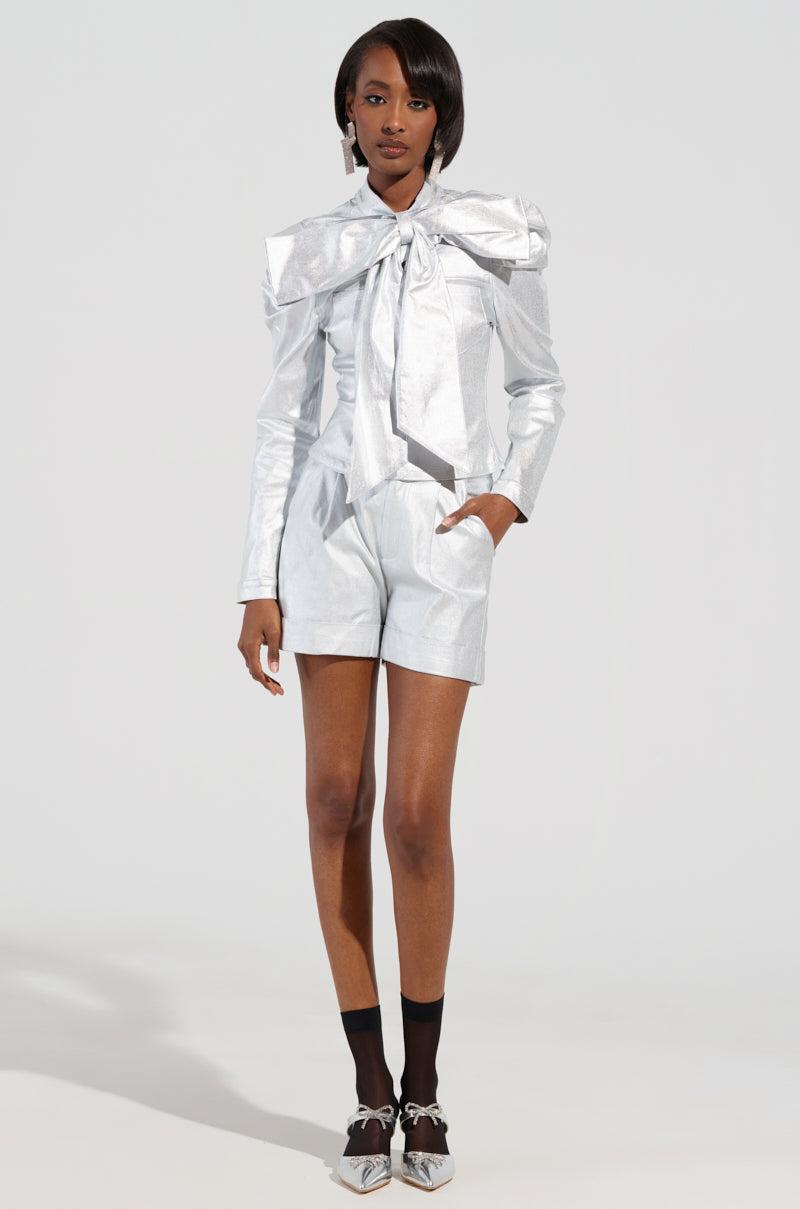 A person stands confidently in the MISS INDEPENDENT METALLIC SHORT IN SILVER, a stylish outfit with metallic shorts adorned with a large bow detail. They have short hair and are posed against a plain background with hands in pockets, complemented by matching socks and shoes. 0