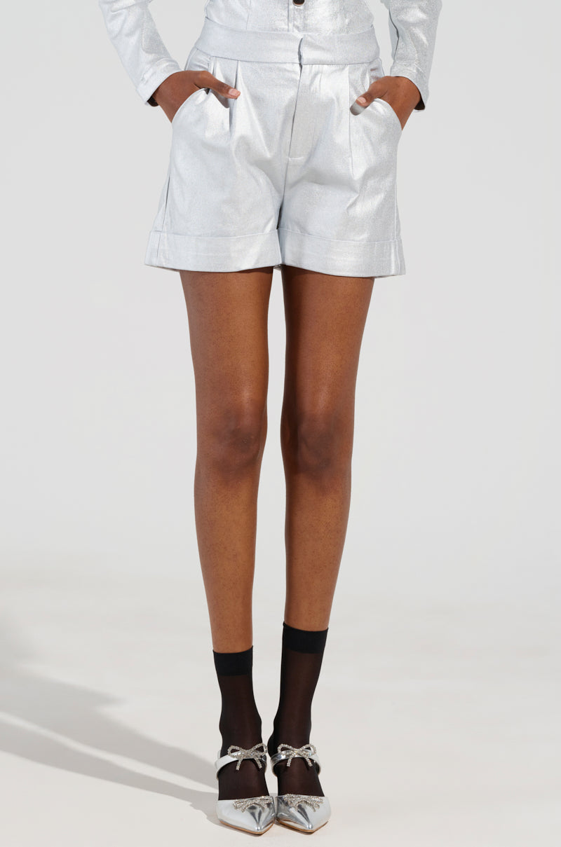 A person dressed in the sleek and stylish MISS INDEPENDENT METALLIC SHORTS IN SILVER, along with a matching top, stands confidently with hands in pockets. Their ensemble is completed with sheer black socks and pointed silver heels adorned with bows, set against a plain light-colored backdrop that accentuates the modern design. 2