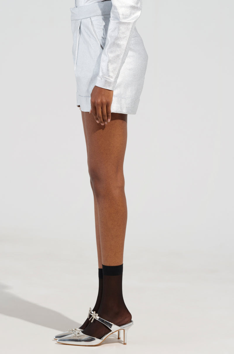 Clad in the MISS INDEPENDENT METALLIC SHORT IN SILVER paired with a long-sleeved top, the individual strikes a pose. Their outfit is completed with metallic pointed heels styled over black ankle socks, set against a stark white backdrop that highlights their daring fashion choice. 3
