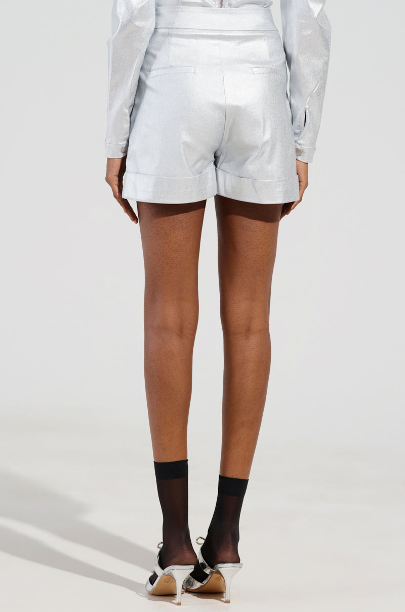 A person stands in the MISS INDEPENDENT METALLIC SHORT IN SILVER and a long-sleeve shirt, facing away. They're wearing black ankle socks and open-toed heels. The background is plain and bright, highlighting the high-rise style of the ensemble. 4
