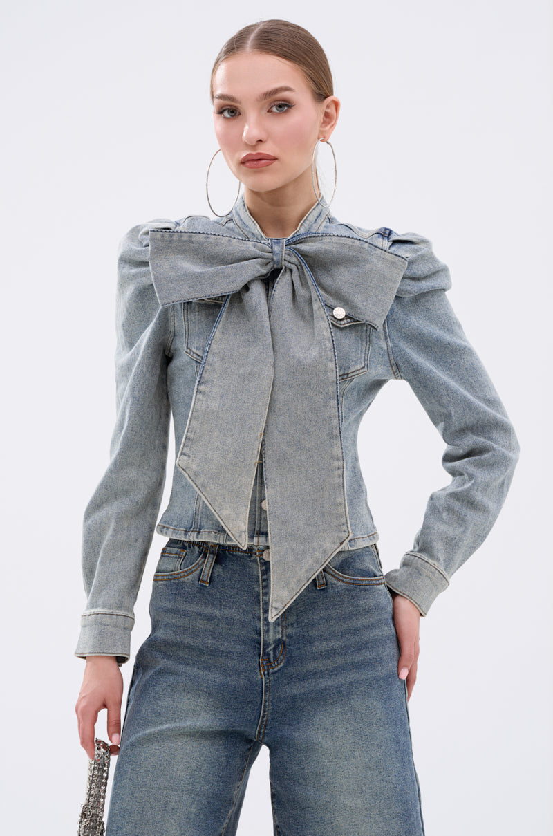 A young woman wears the WAIT FOR U DENIM BOW BLOUSE TOP with puff shoulders and a statement collar bow, styled with high-waisted blue jeans, hoop earrings, and straight hair pulled back, set against a plain white background.