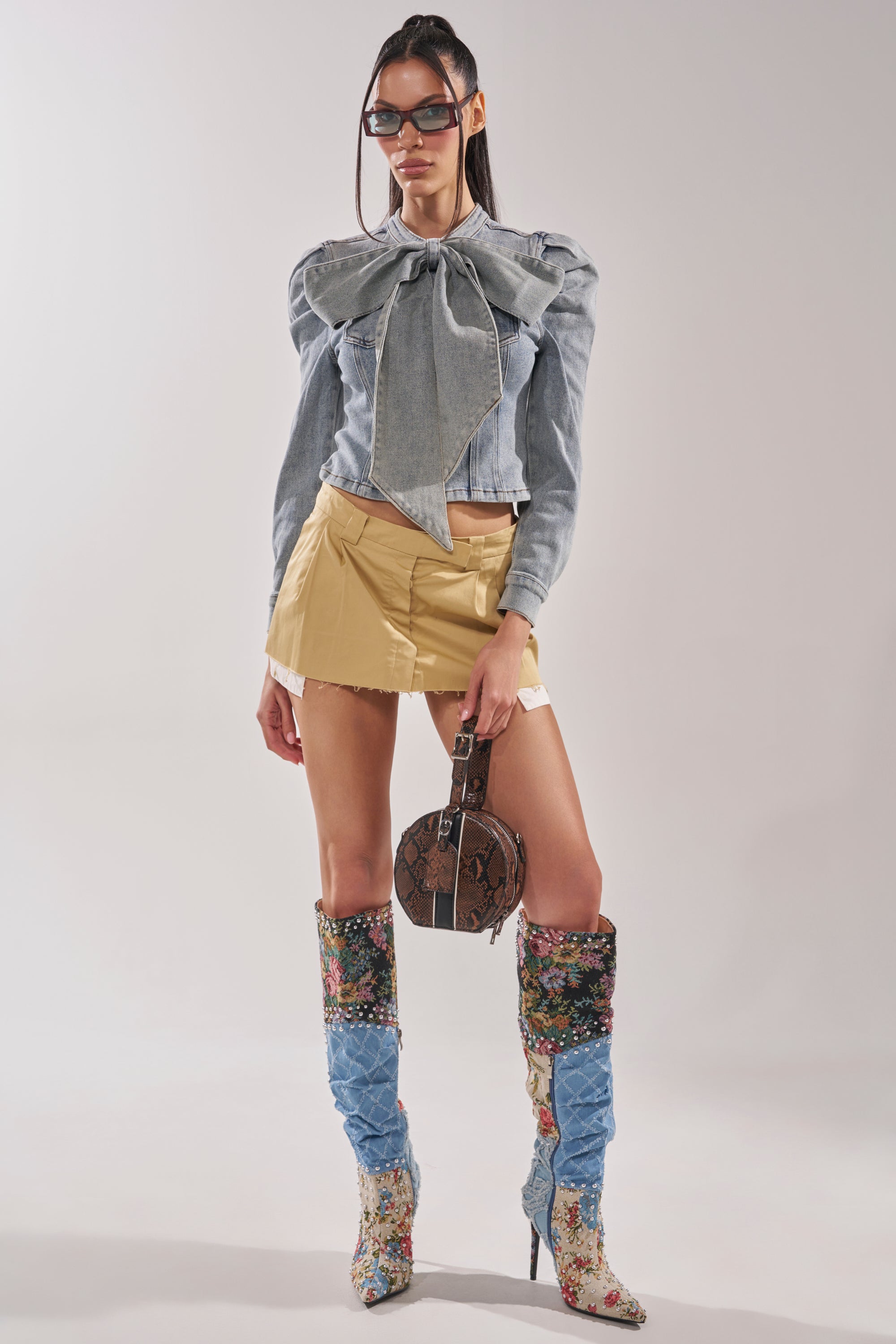 A woman in the WAIT FOR U DENIM BOW BLOUSE TOP, gold mini skirt, knee-high floral and denim boots, and sunglasses holds a round snakeskin handbag, posing confidently against a plain background. 9