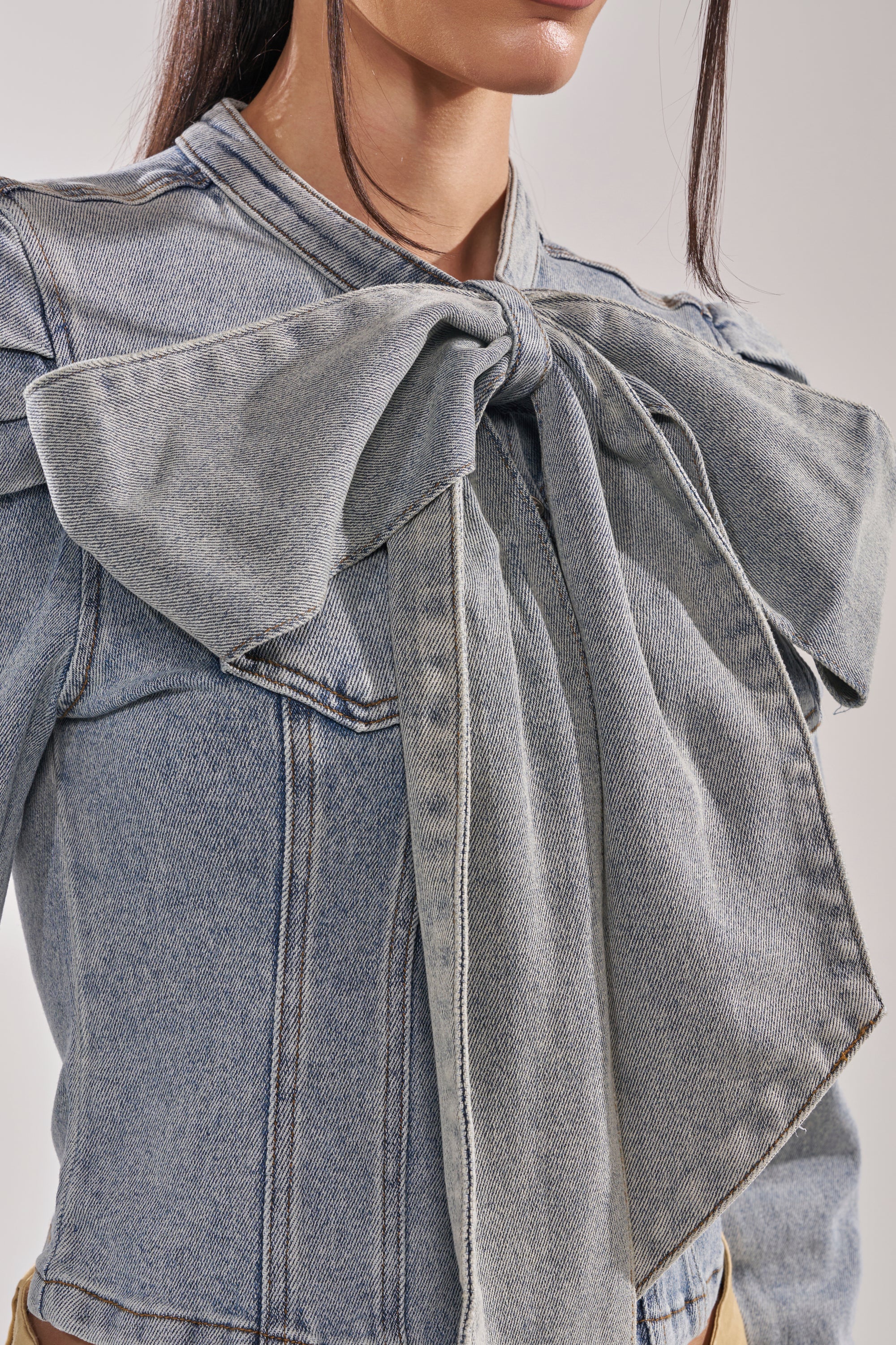 A woman models the WAIT FOR U DENIM BOW BLOUSE TOP by AKIRA Label, featuring a light wash and a large statement bow at the collar; the image focuses on her upper torso with her face partially out of frame. 10