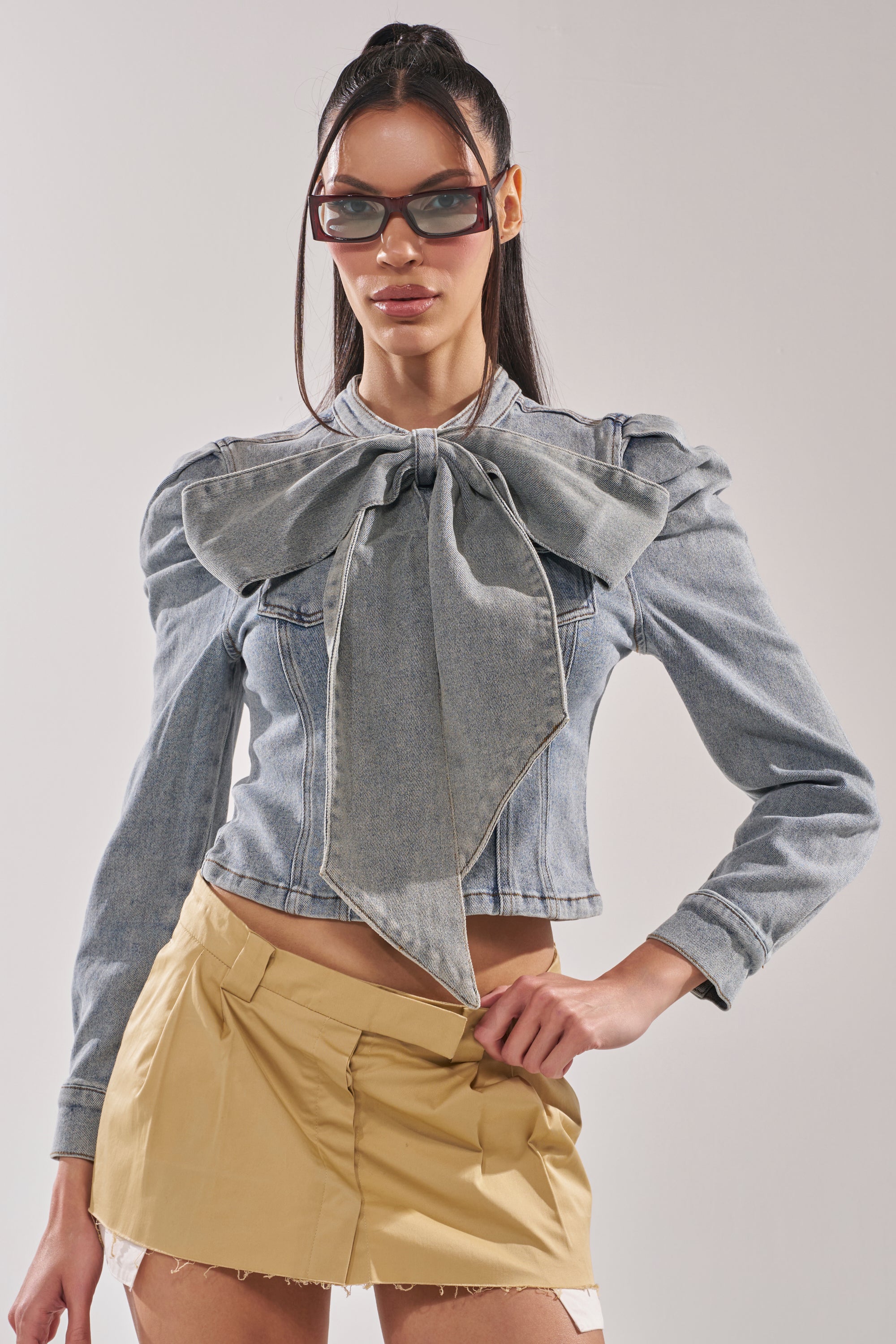 A woman stands confidently with one hand on her hip, wearing large sunglasses, the WAIT FOR U DENIM BOW BLOUSE TOP with puffed shoulders, and a beige mini skirt against a plain background. 0