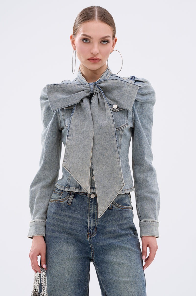 A woman wearing the WAIT FOR U DENIM BOW BLOUSE TOP with puff shoulders and matching high-waisted denim jeans stands against a plain white background, holding a small purse.