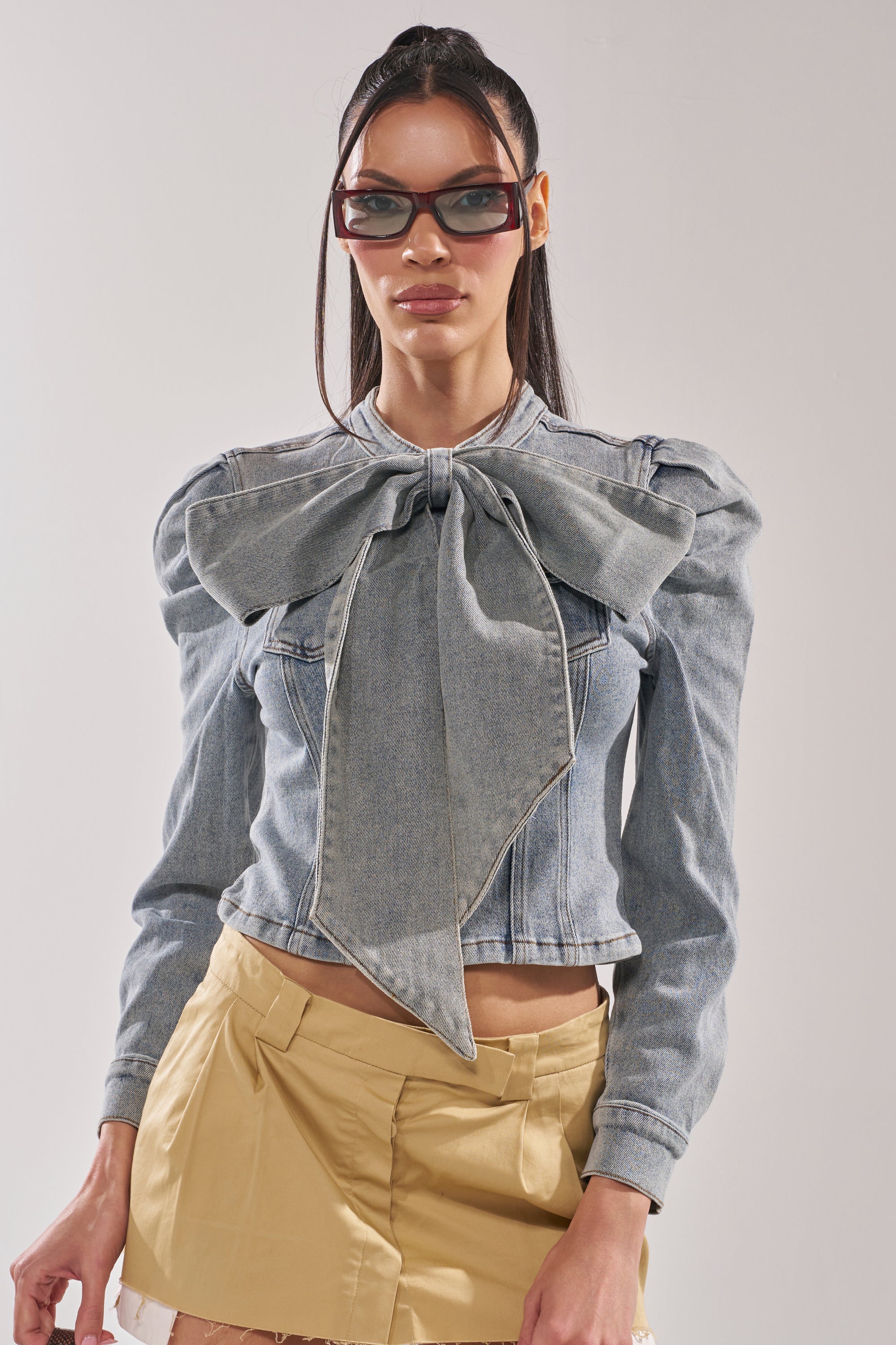 A woman wearing sunglasses, the WAIT FOR U DENIM BOW BLOUSE TOP with puffed shoulders and a large bow at the collar, and a beige mini skirt stands against a plain light background. 1