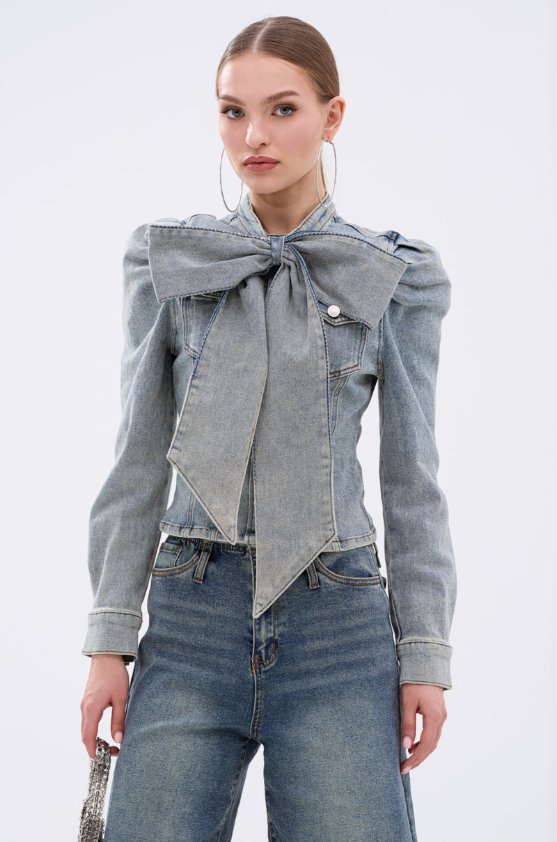 A woman wears the WAIT FOR U DENIM BOW BLOUSE TOP with puff shoulders and high-waisted jeans, standing against a plain white background. Her straight hair is pulled back, and she carries a small shiny purse.