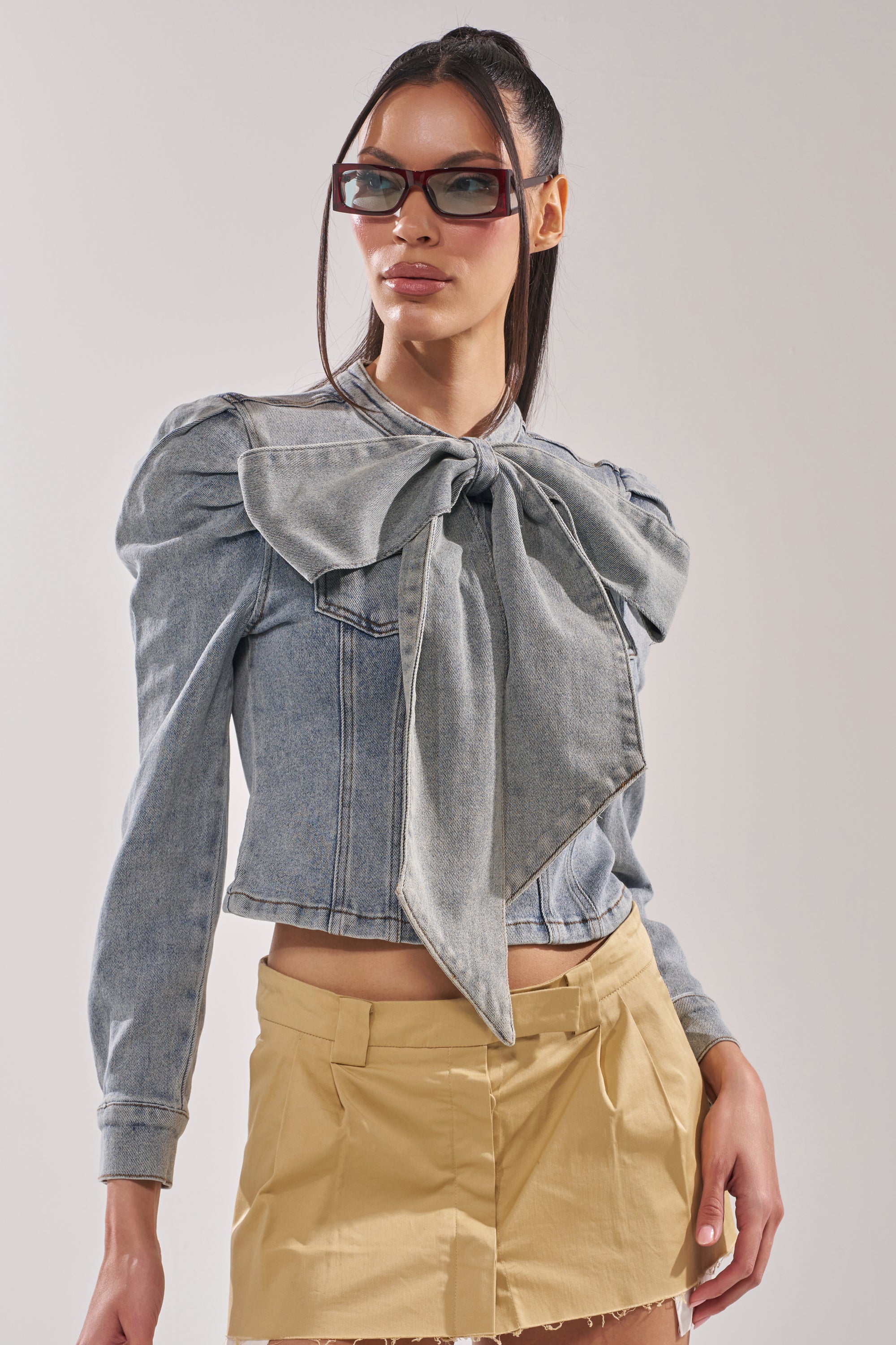 A woman confidently poses against a plain background in the WAIT FOR U DENIM BOW BLOUSE TOP with puffed shoulders, paired with a beige pleated mini skirt and rectangular sunglasses. 2