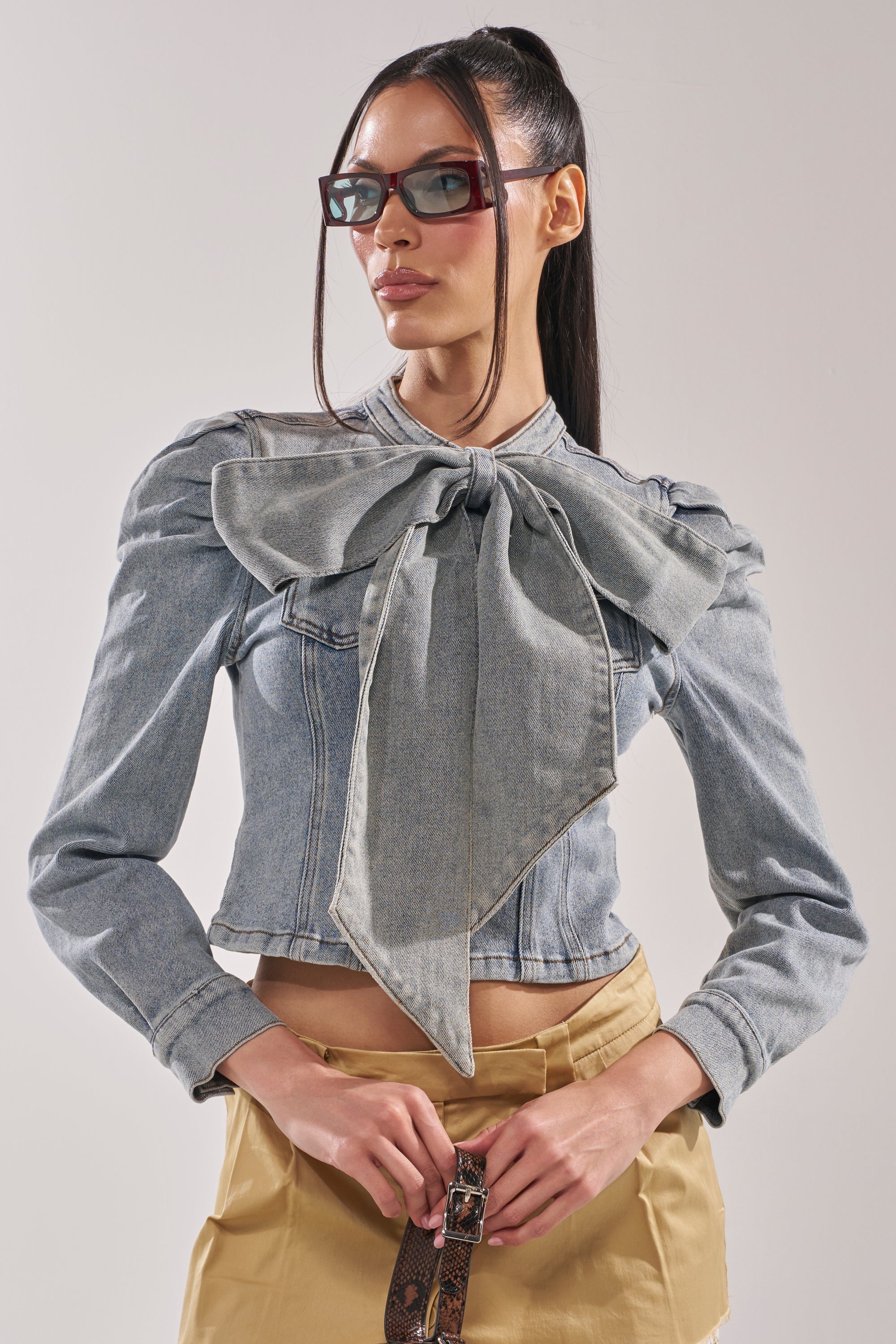 A woman wears the WAIT FOR U DENIM BOW BLOUSE TOP with puffed shoulders, beige high-waisted pants, sunglasses, and a high ponytail, standing against a plain light background. 3