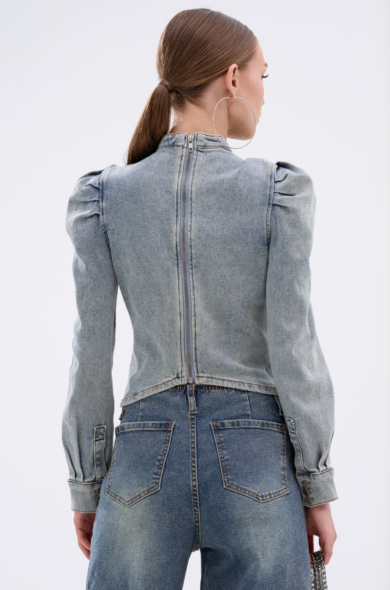 A woman with straight brown hair in a low ponytail wears the WAIT FOR U DENIM BOW BLOUSE TOP, a light blue denim jacket with puff shoulders and an exposed back zipper, paired with high-waisted jeans. She faces away from the camera.