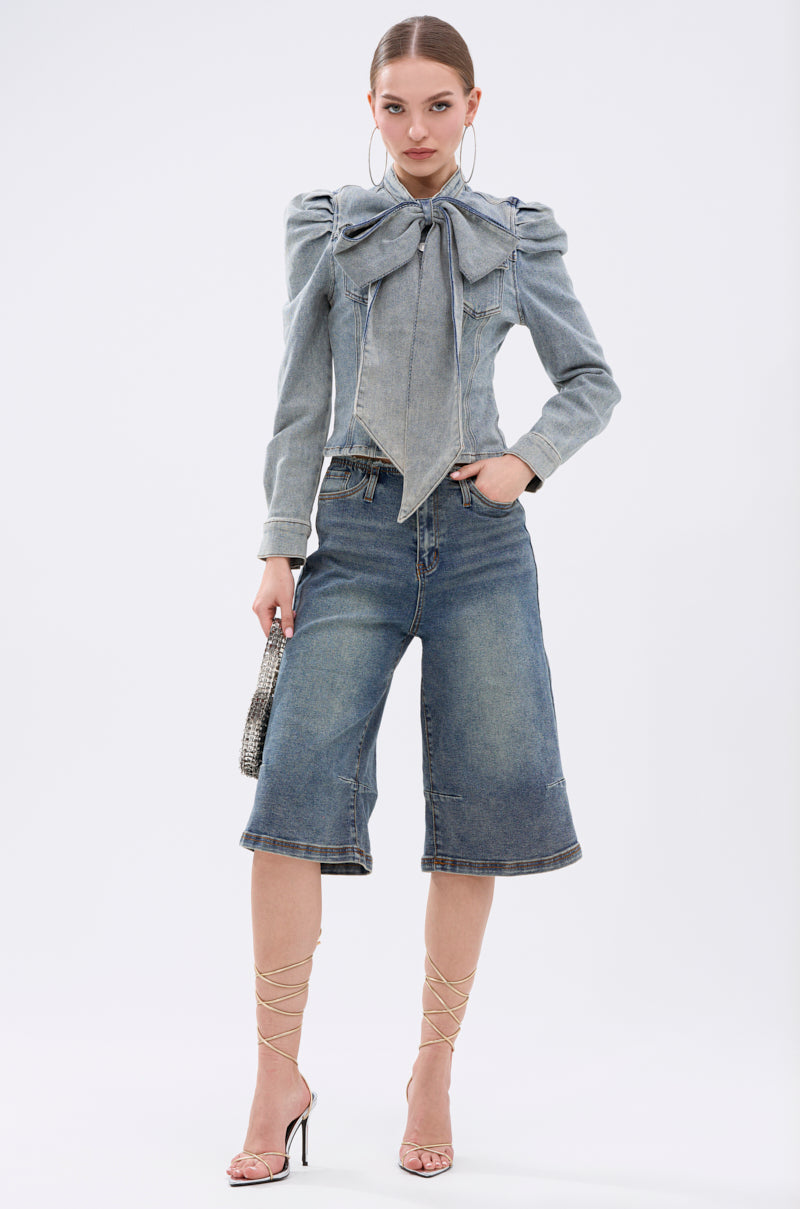 A woman poses in the WAIT FOR U DENIM BOW BLOUSE TOP with puff shoulders, knee-length denim shorts, lace-up high-heeled sandals, large hoop earrings, and a small handbag against a plain white background.
