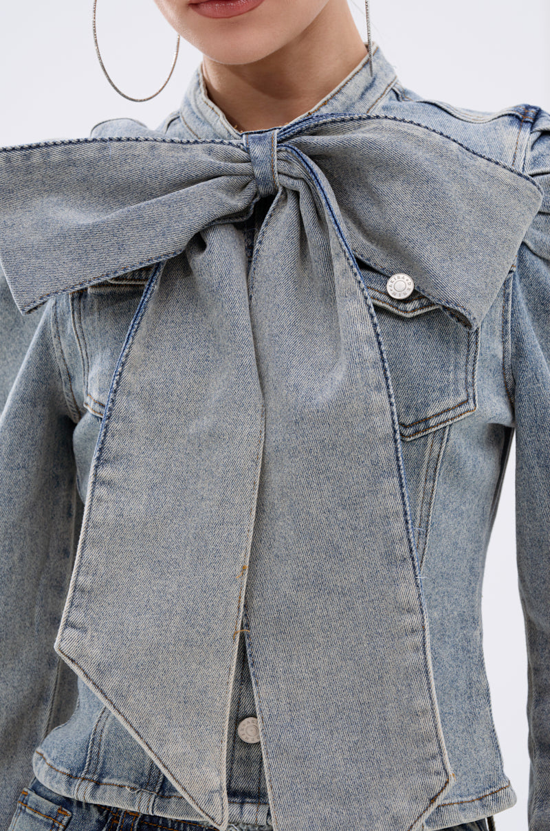 Seen partially, a person wears the WAIT FOR U DENIM BOW BLOUSE TOP, featuring a striking oversized denim bow at the collar and subtle puff shoulders; only their lower face is visible.