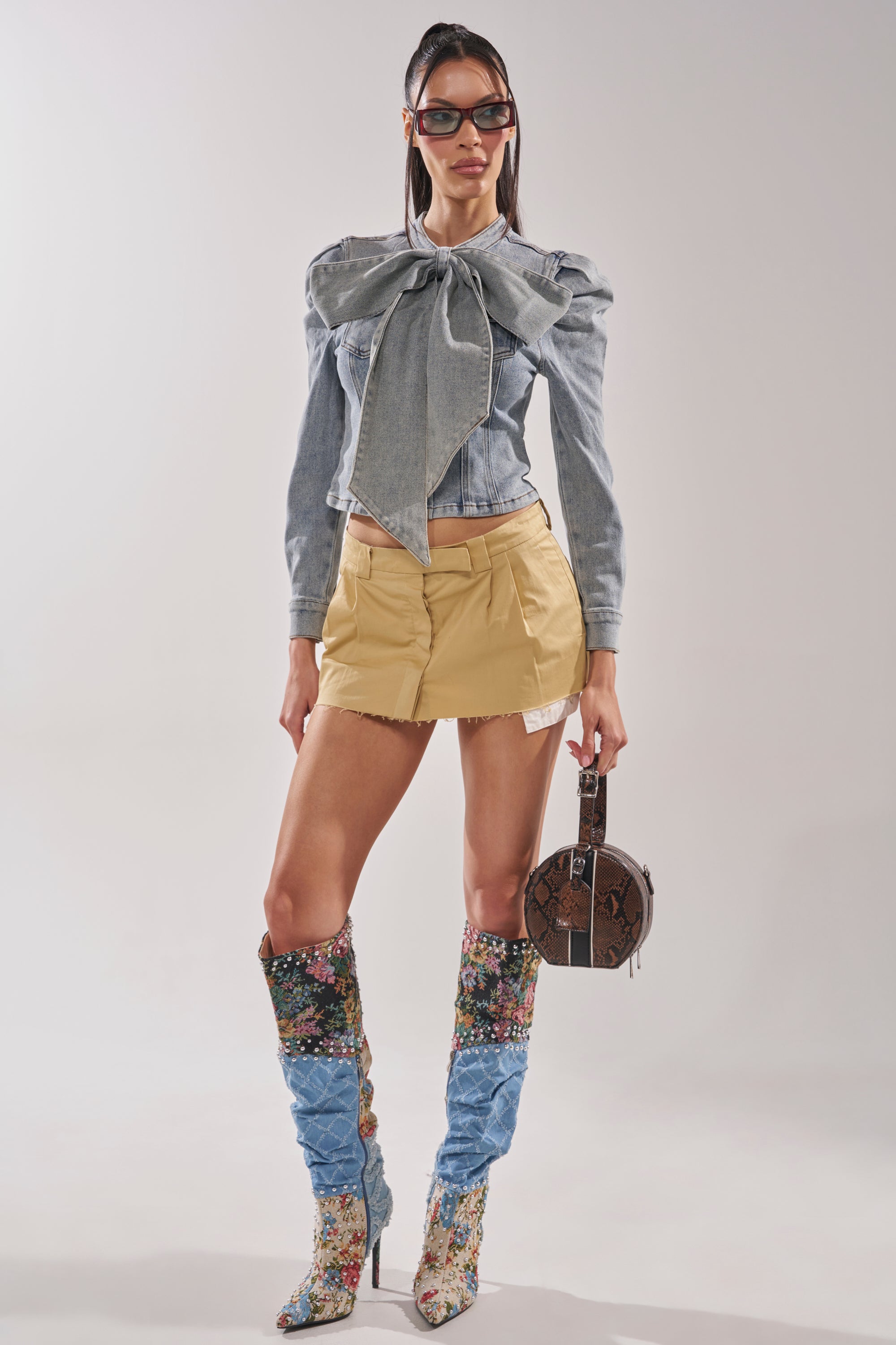 A woman stands against a plain light background wearing the WAIT FOR U DENIM BOW BLOUSE TOP with puffed shoulders, a short beige skirt, floral and denim knee-high boots, sunglasses, and holding a round brown handbag. 7