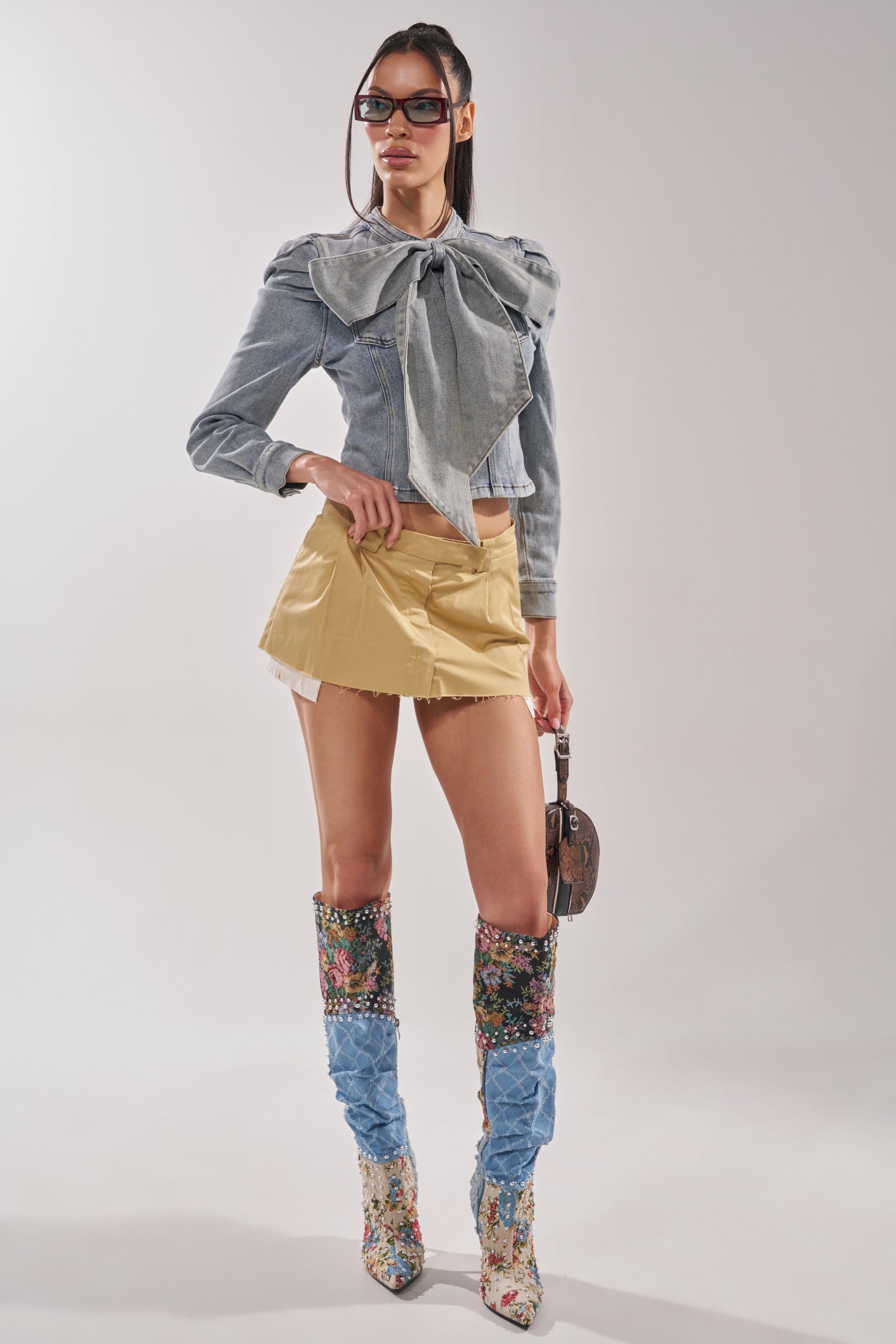 A woman models the WAIT FOR U DENIM BOW BLOUSE TOP with puffed shoulders, paired with a short tan skirt, tall blue-accented floral boots, sunglasses, and a small round purse against a plain background. 8