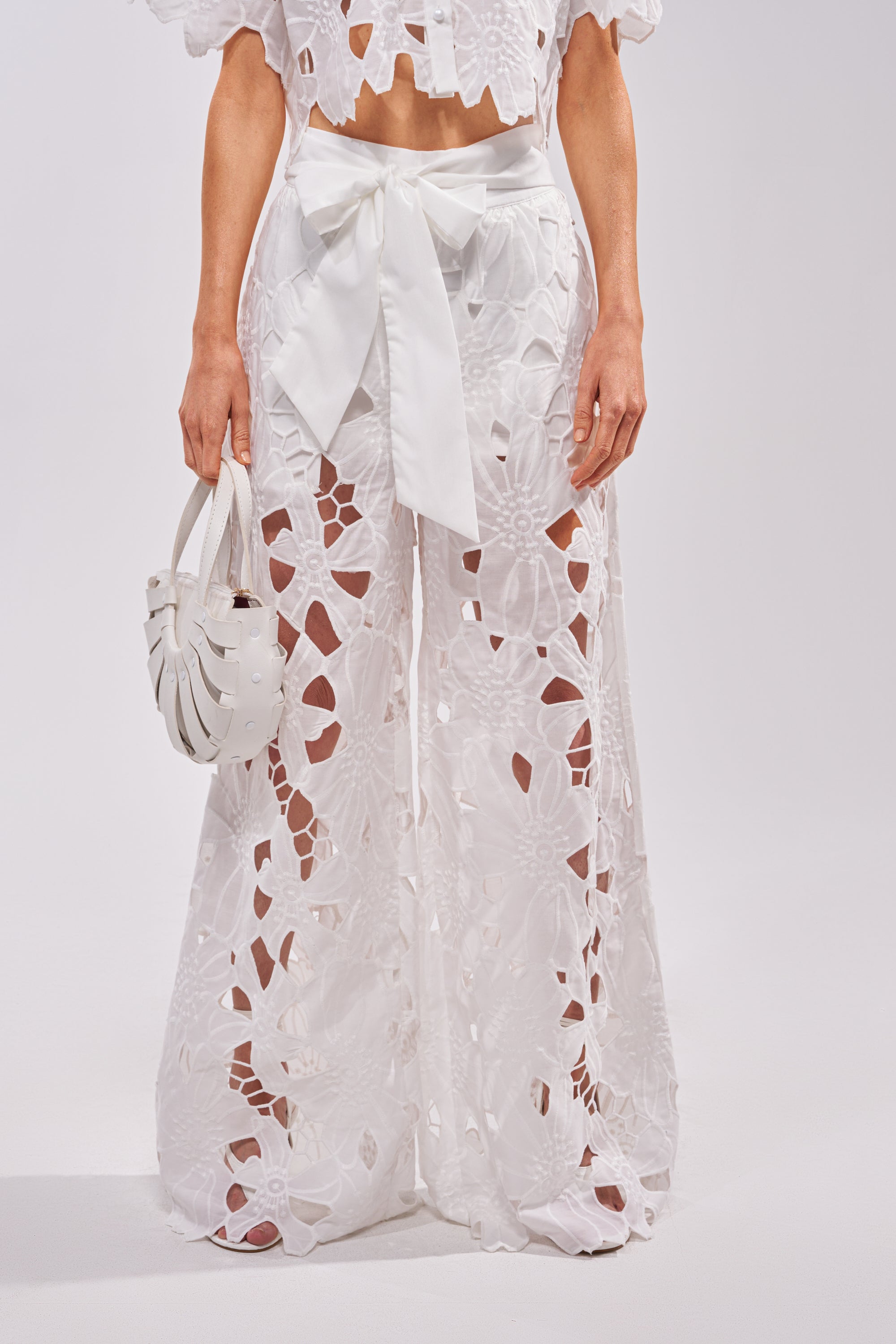Wearing the DOLLY FLORAL PALAZZO PANT IN WHITE, which features a high waist and large floral cut-outs, paired with an oversized front bow and a small white handbag. The top is partially visible. 0