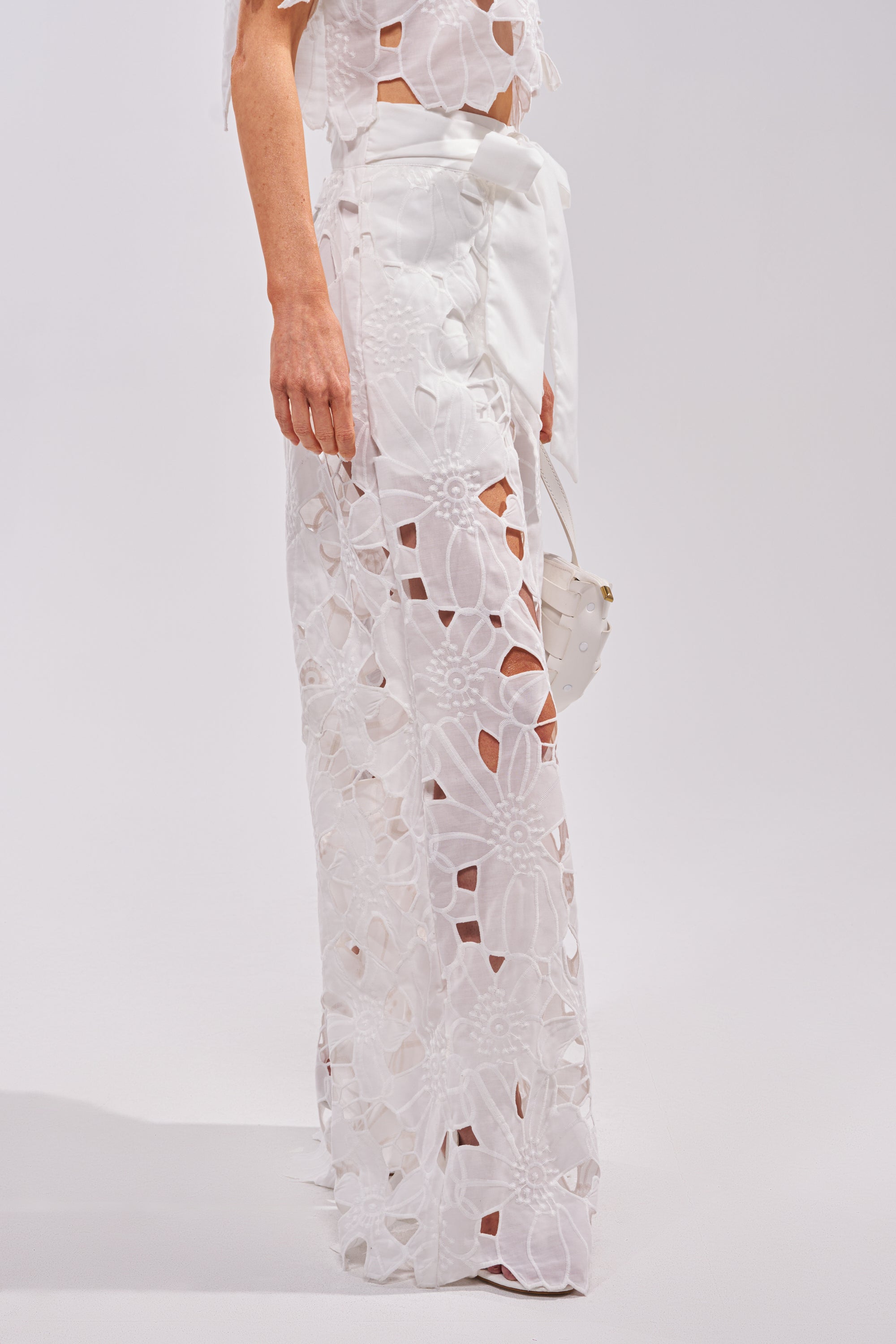 Wearing the DOLLY FLORAL PALAZZO PANT IN WHITE with floral cutouts and a matching top, she holds a small white bag while standing against a plain light background. 1