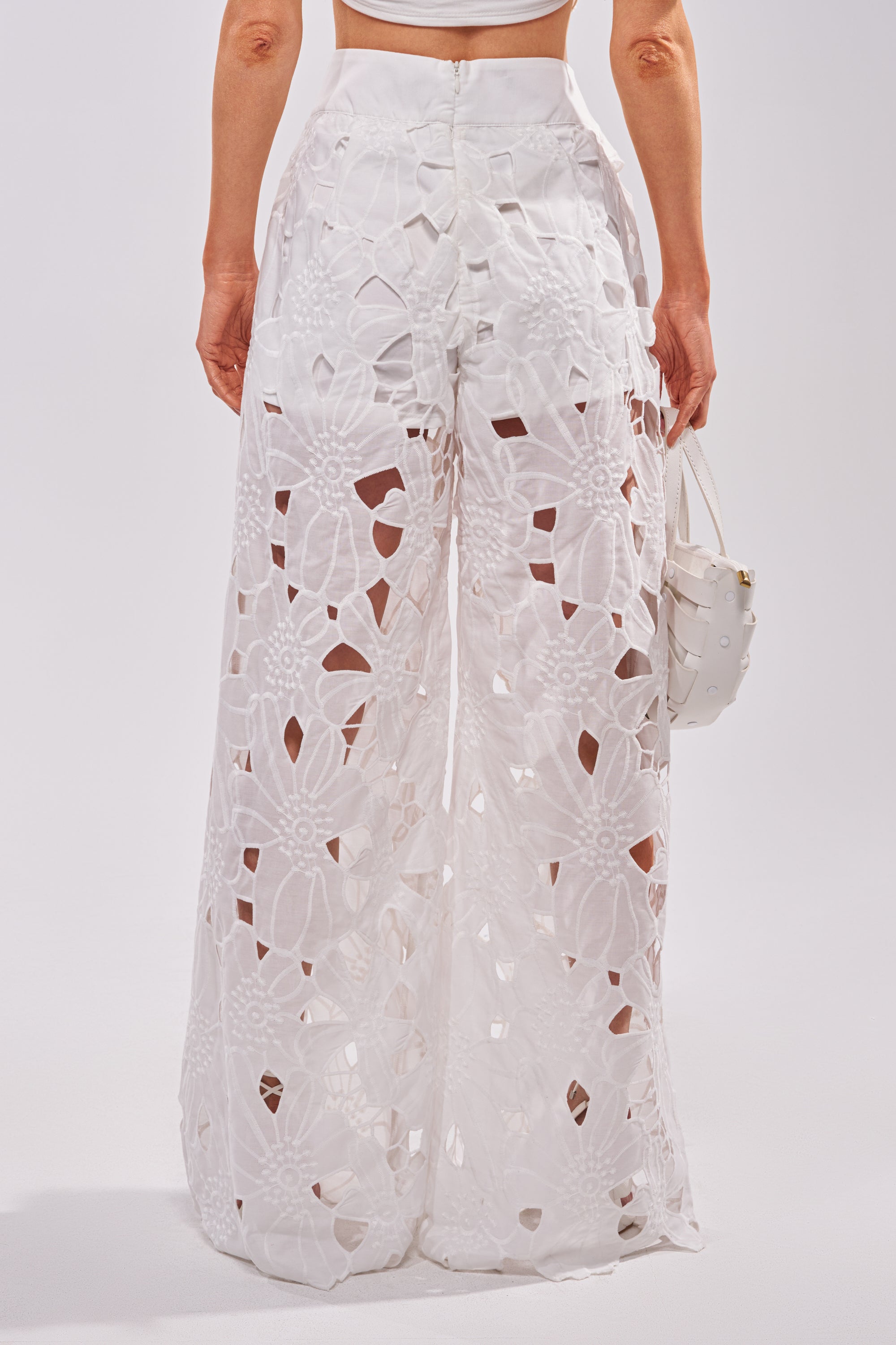 A person shown from the back against a plain background, wearing the DOLLY FLORAL PALAZZO PANT IN WHITE—high-waisted with large floral lace cut-outs—and carrying a small white handbag, creating an elegant summer look. 2
