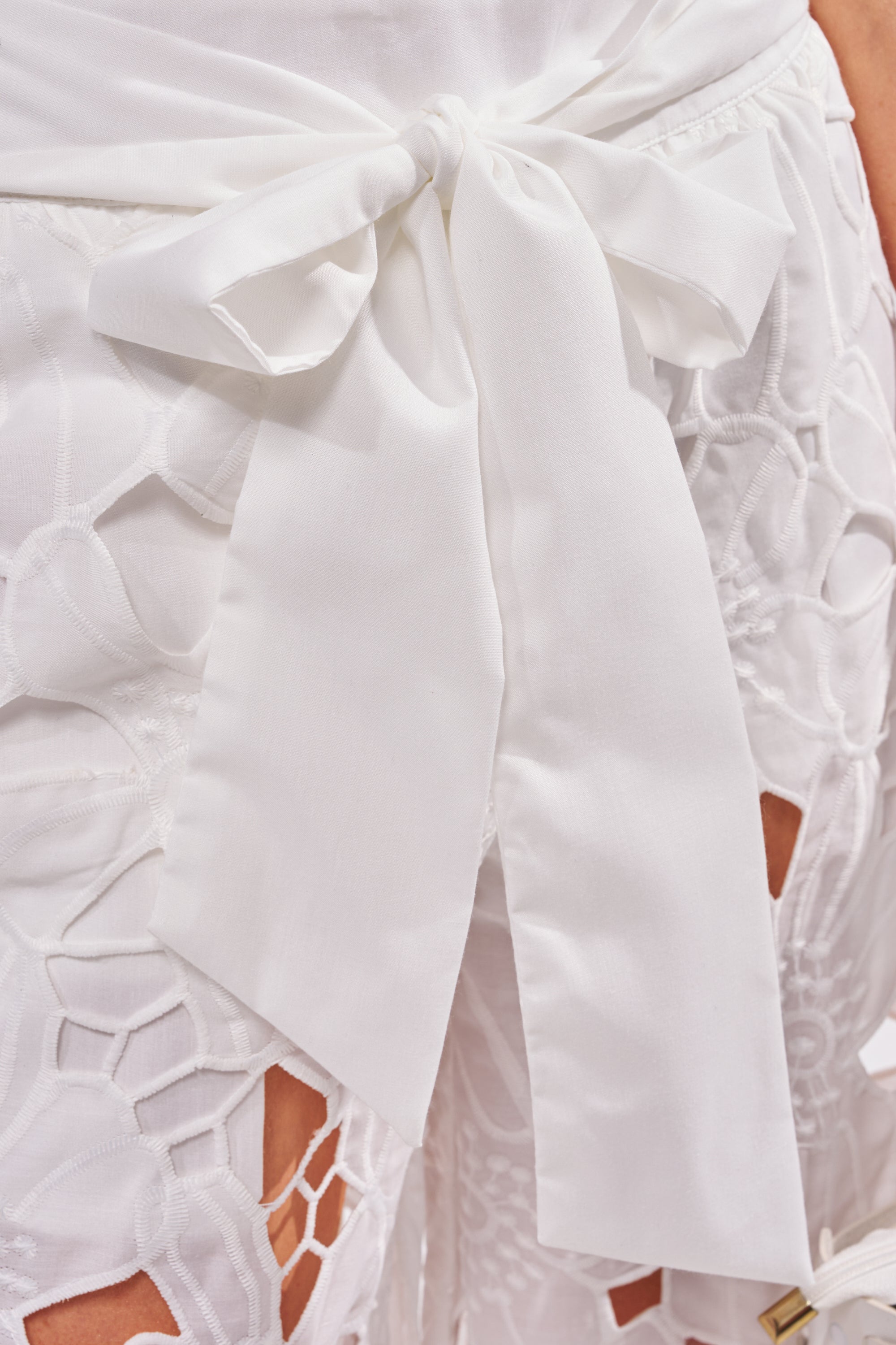 Close-up of a person wearing the DOLLY FLORAL PALAZZO PANT IN WHITE, featuring white lace fabric with geometric patterns and a large high-waisted white fabric bow. 5
