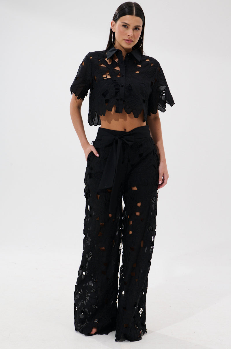 A woman poses against a plain background in the DOLLY FLORAL PALAZZO PANT set, featuring a black short-sleeve button-up crop top and matching high-waisted floral palazzo pants with lace cutouts, wide legs, and a sash belt.
