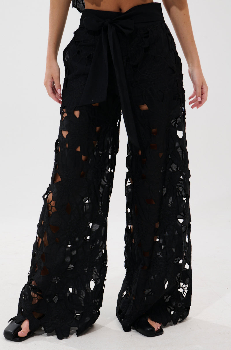 A person wears the DOLLY FLORAL PALAZZO PANT—embroidered lace, wide-leg with tie belt and floral cut-outs—paired with open-toe black shoes. The sheer fabric elegantly showcases the legs underneath.