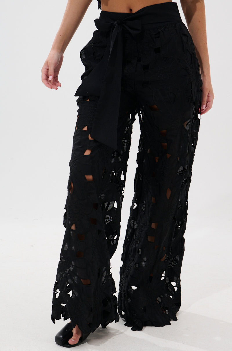 A person wearing the DOLLY FLORAL PALAZZO PANT—high-waisted, wide-leg pants with embroidered lace, large cut-out patterns, and a fabric belt—paired with open-toe shoes, stands against a plain white background.
