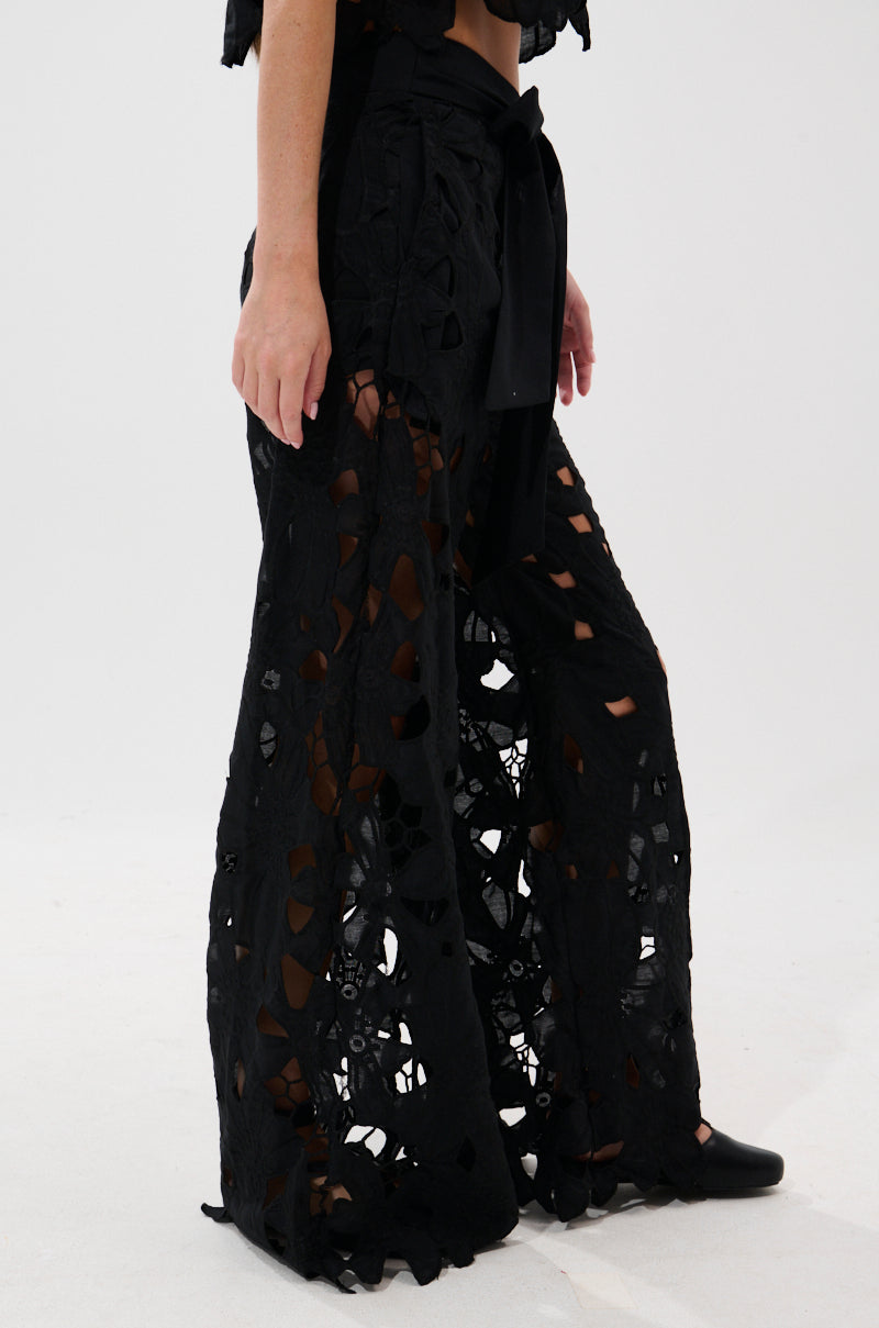 Wearing the DOLLY FLORAL PALAZZO PANT, a person showcases high-waisted black lace pants with wide legs, floral embroidery, and cut-out details. The look is finished with a matching fabric belt against a light background.