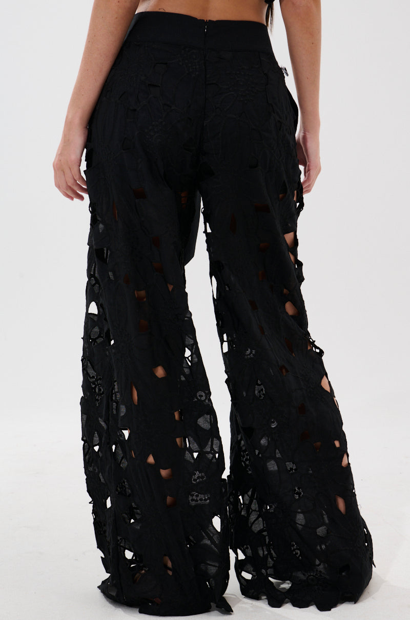 A woman is seen from the back wearing the DOLLY FLORAL PALAZZO PANT, high-waisted with embroidered lace, sheer cutout detailing, a wide-leg silhouette, zip closure at the back, and an all-over floral pattern.