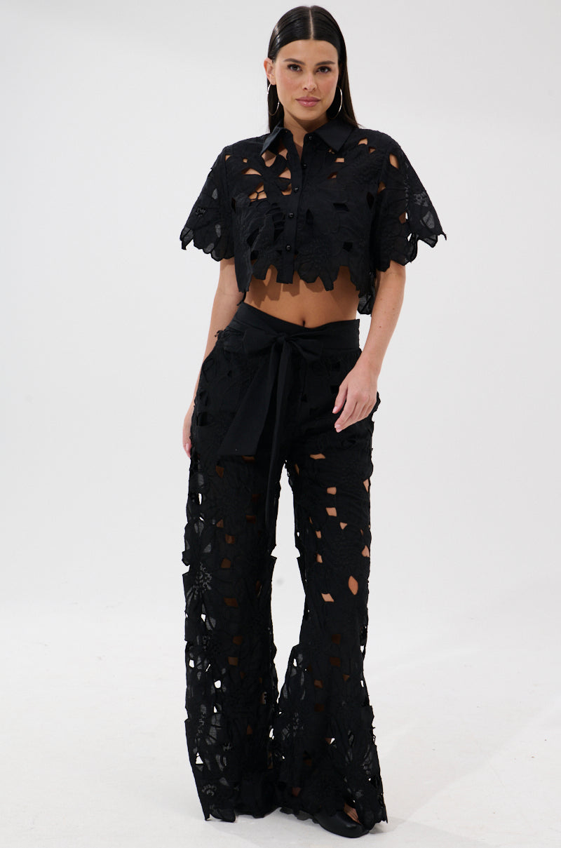 A woman with long dark hair poses against a plain white background, wearing the DOLLY FLORAL PALAZZO PANT—a black, wide-leg palazzo pant with floral cutout patterns—paired with a matching short-sleeve, button-up crop top.