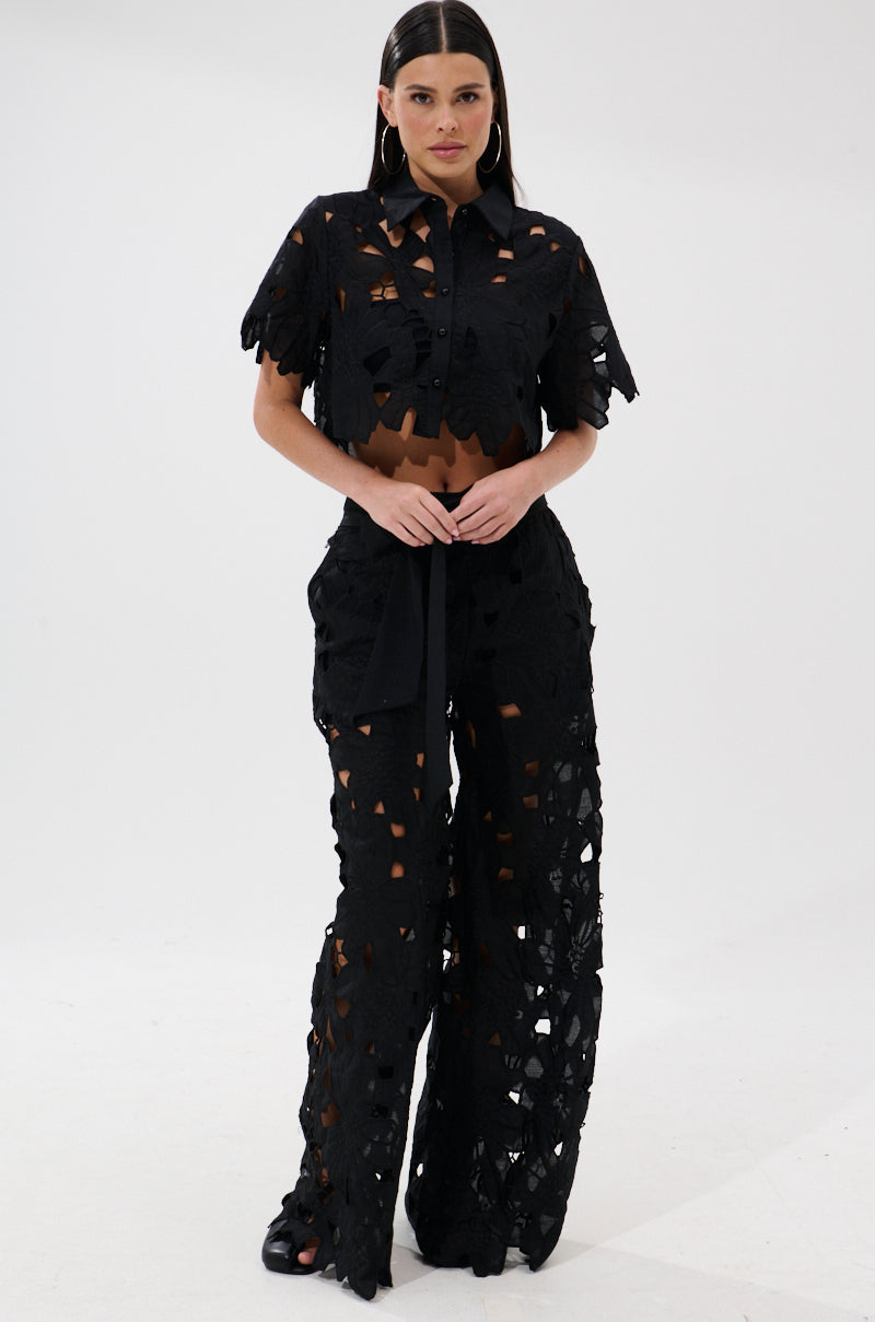 A woman faces forward wearing the DOLLY FLORAL PALAZZO PANT—high-waisted, embroidered lace pants with cutout patterns—paired with a black, short-sleeve, button-up cropped top. She has long straight hair, hoop earrings, and sandals.