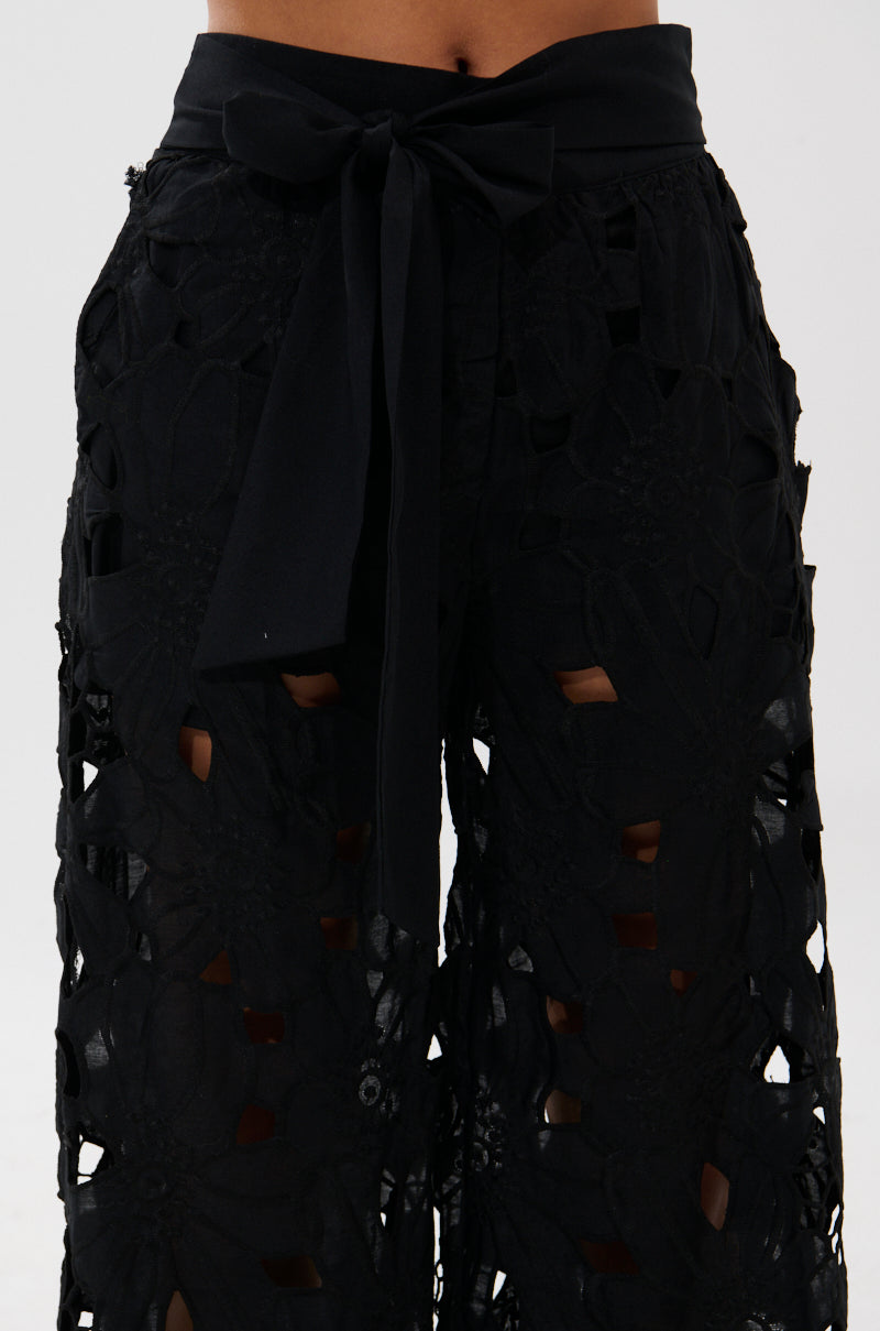 Close-up of a person wearing the DOLLY FLORAL PALAZZO PANT, featuring black embroidered lace with a floral pattern, sheer cutouts, a wide-leg silhouette, and a fabric belt tied in a bow. The background is plain and light-colored.